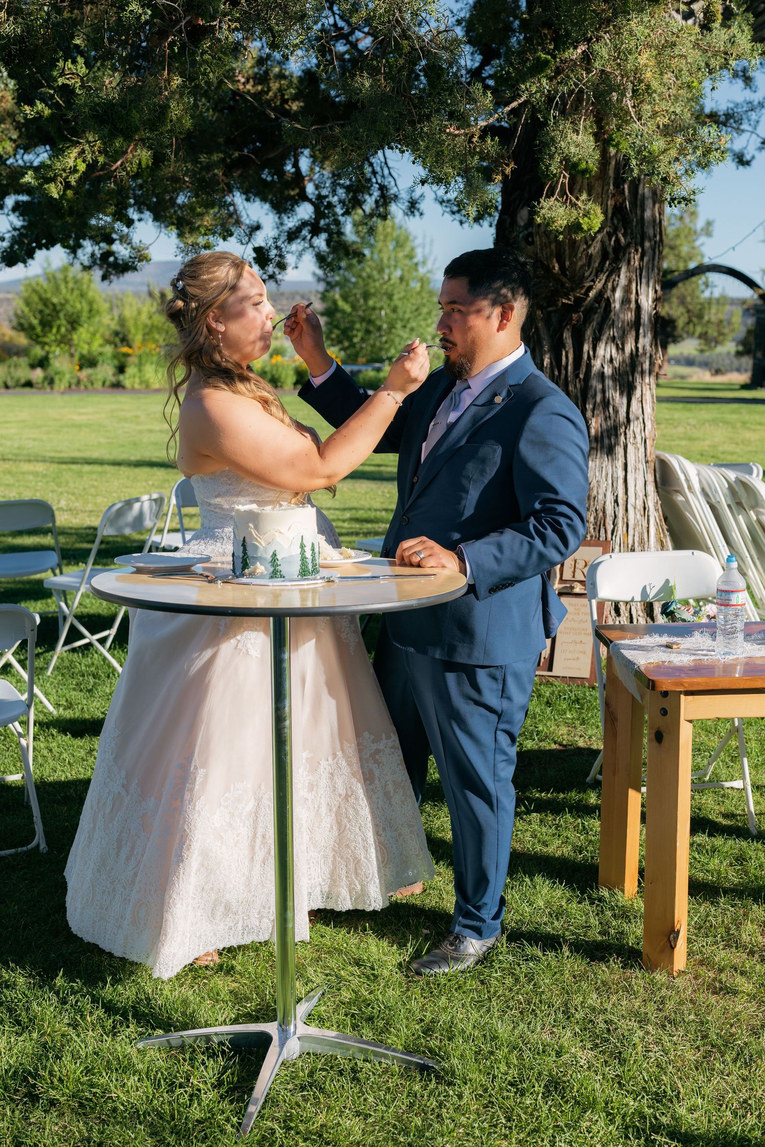 Bride and groom share cake cutting moment at outdoor wedding reception on green lawn.