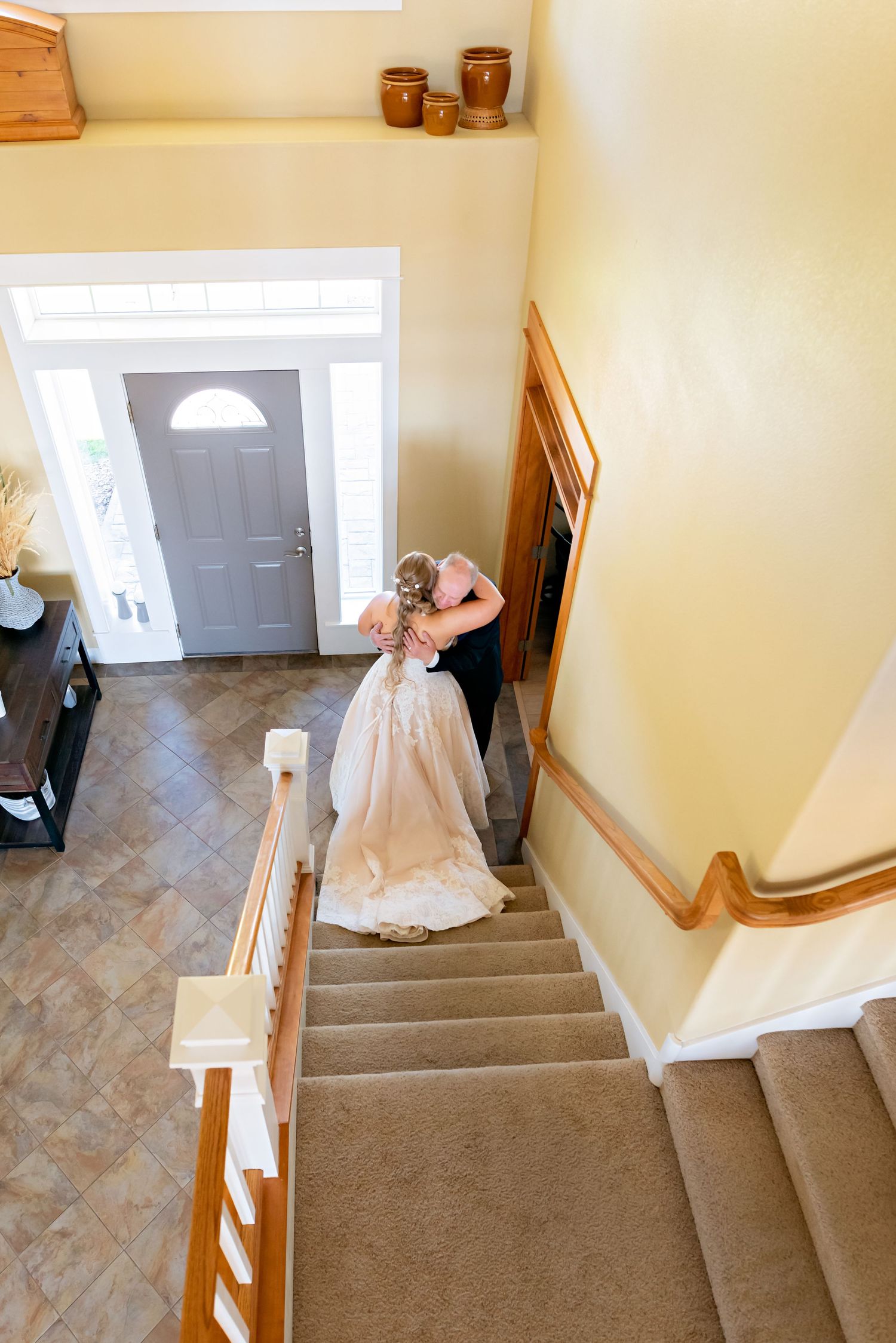 Aerial view of bride in white gown being helped by bridesmaid at bottom of carpeted staircase near front door.