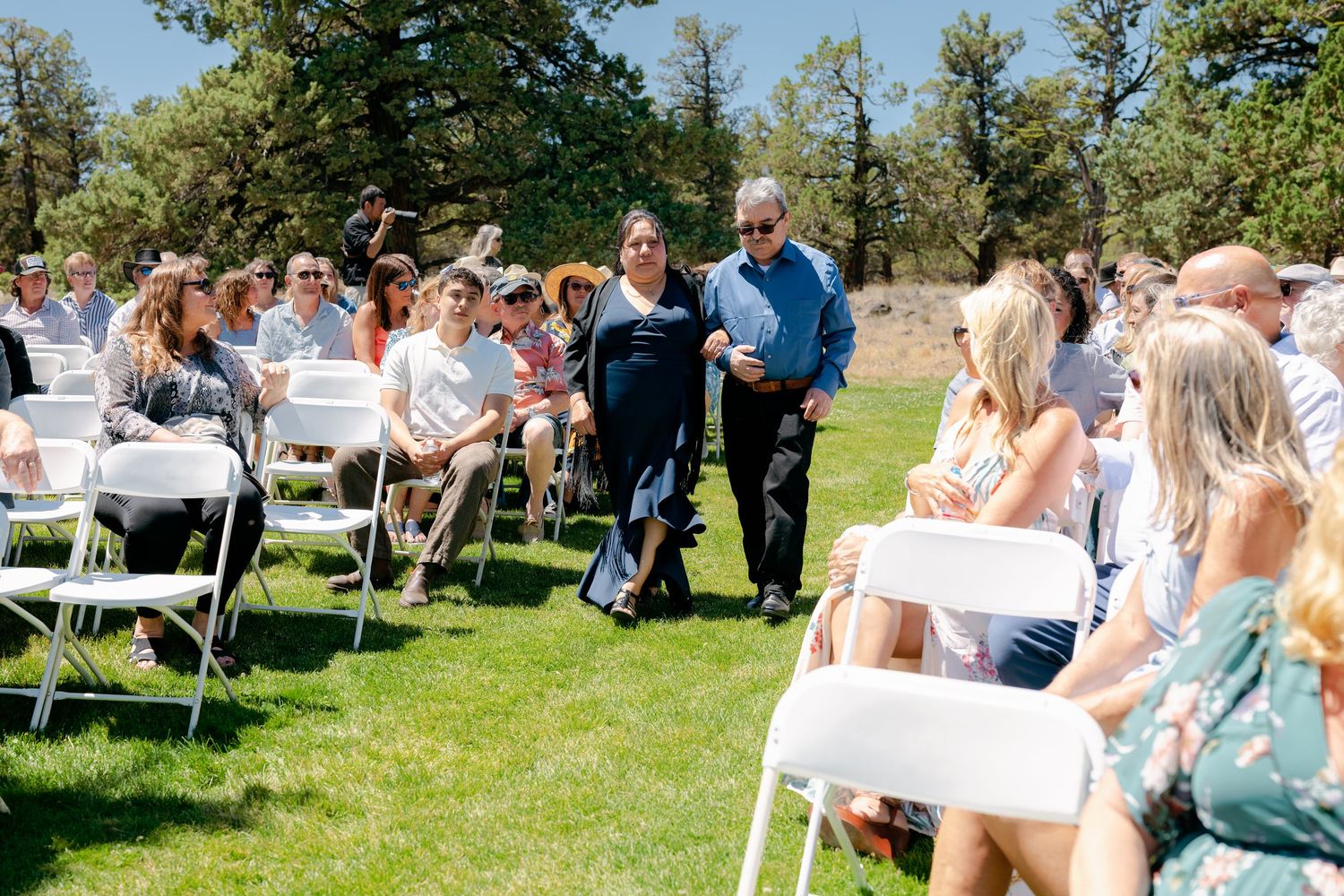 Wedding guests seated in white chairs on green lawn as couple walks down outdoor ceremony aisle.