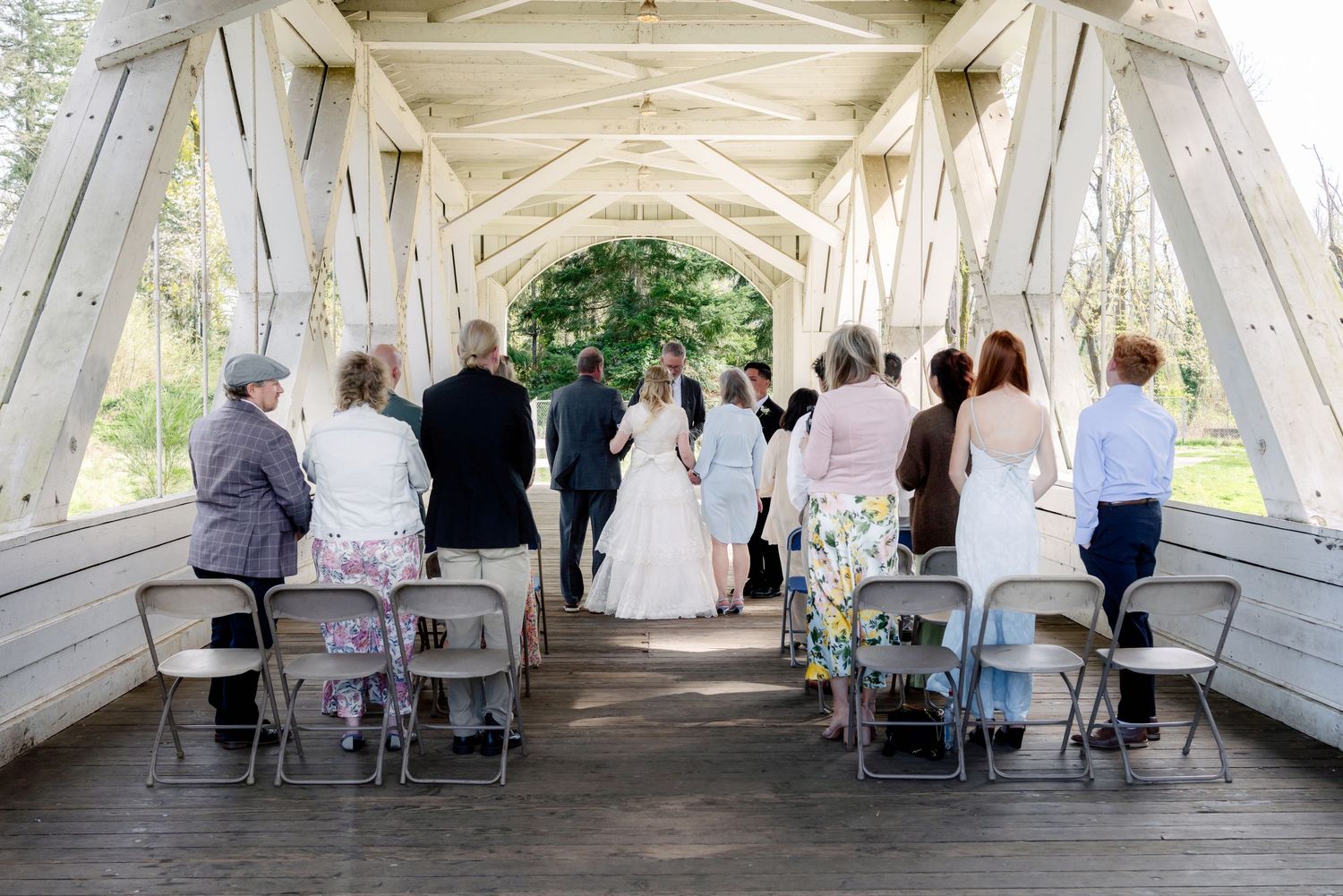 Wedding ceremony inside a white covered bridge with guests seated in folding chairs.