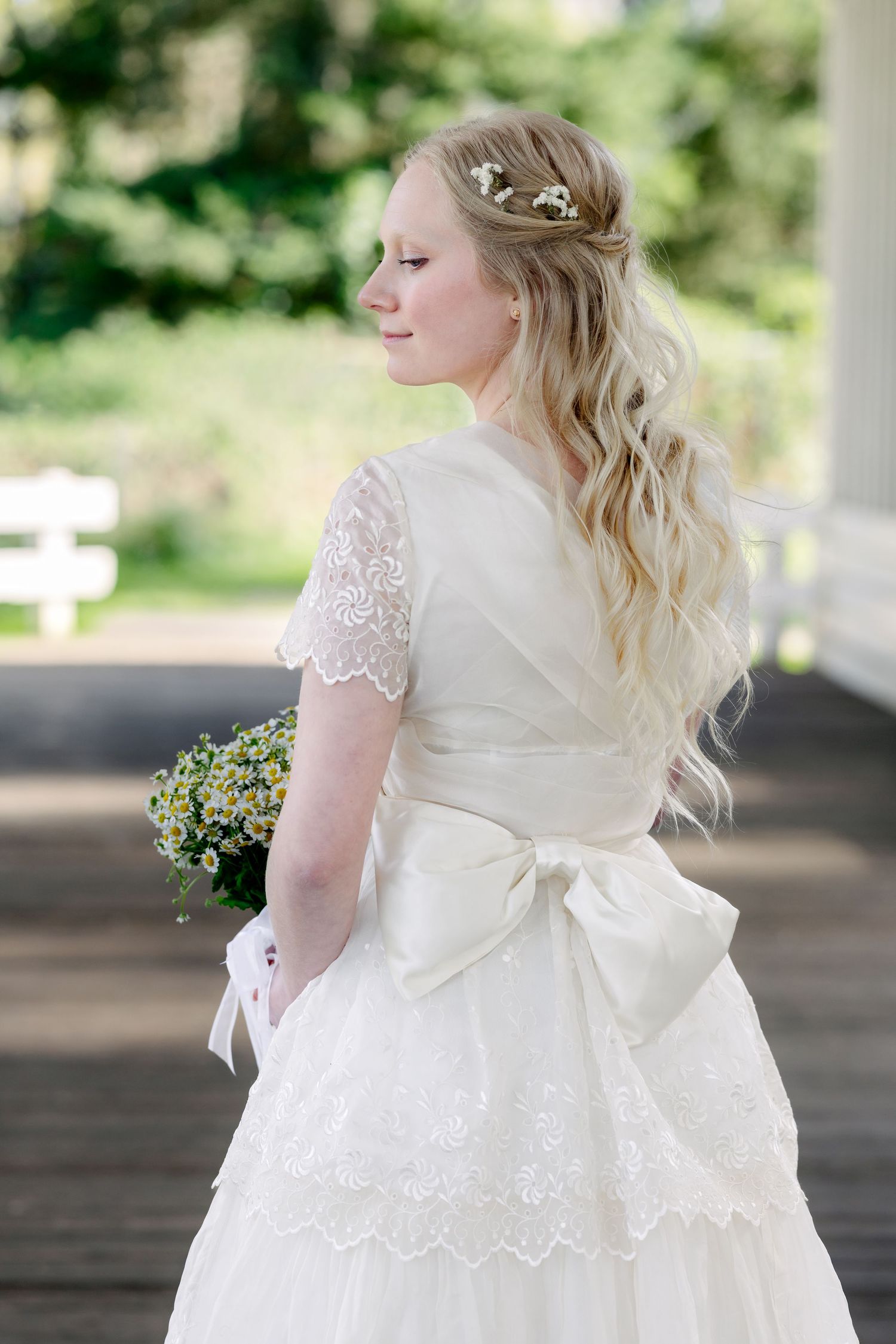 Blonde bride in white lace wedding dress with satin bow holds wildflower bouquet, hair adorned with small flowers.