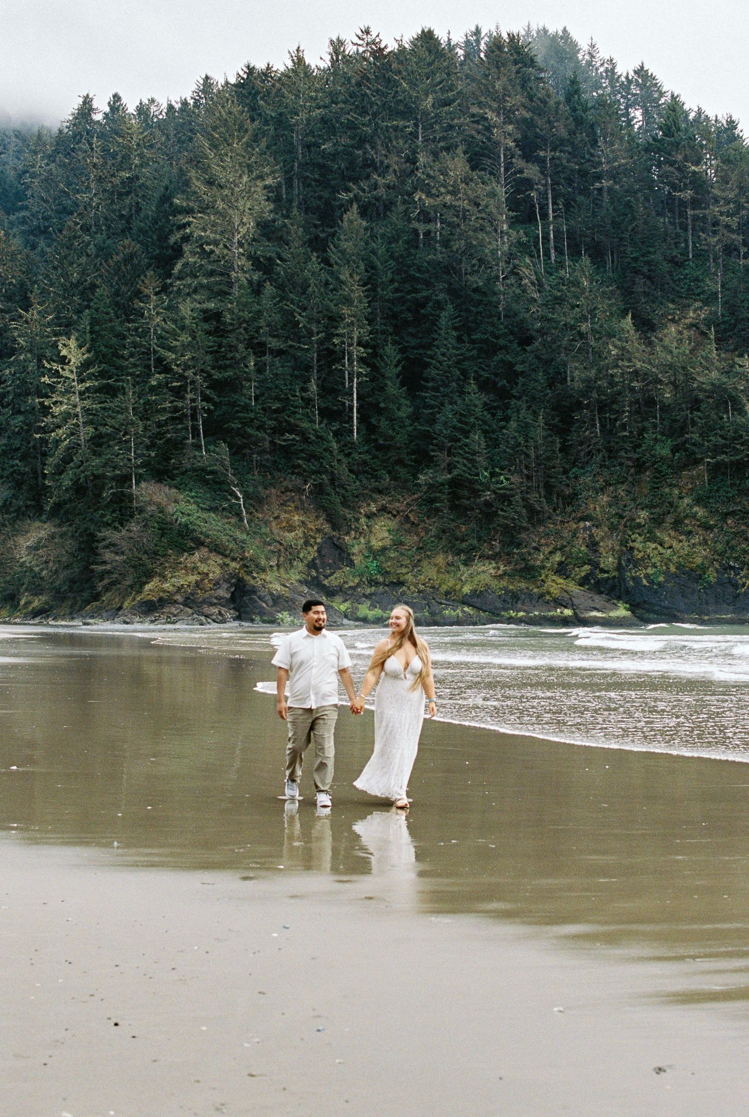 Couple in white outfits holding hands and walking along a scenic beach with dense evergreen forest backdrop.
