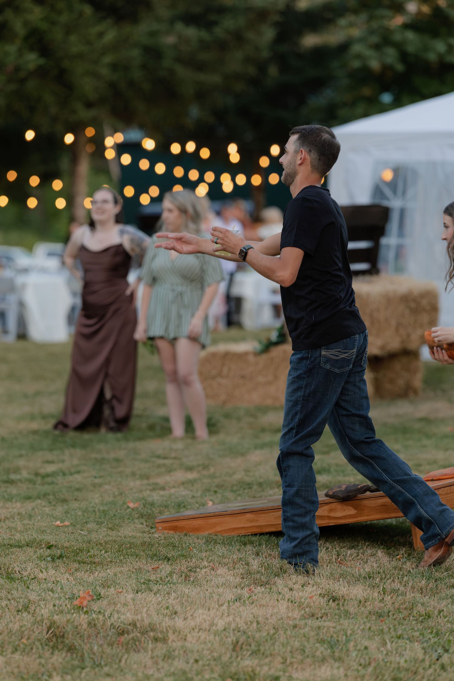 A man plays cornhole at an outdoor wedding reception with string lights and hay bales in the background.