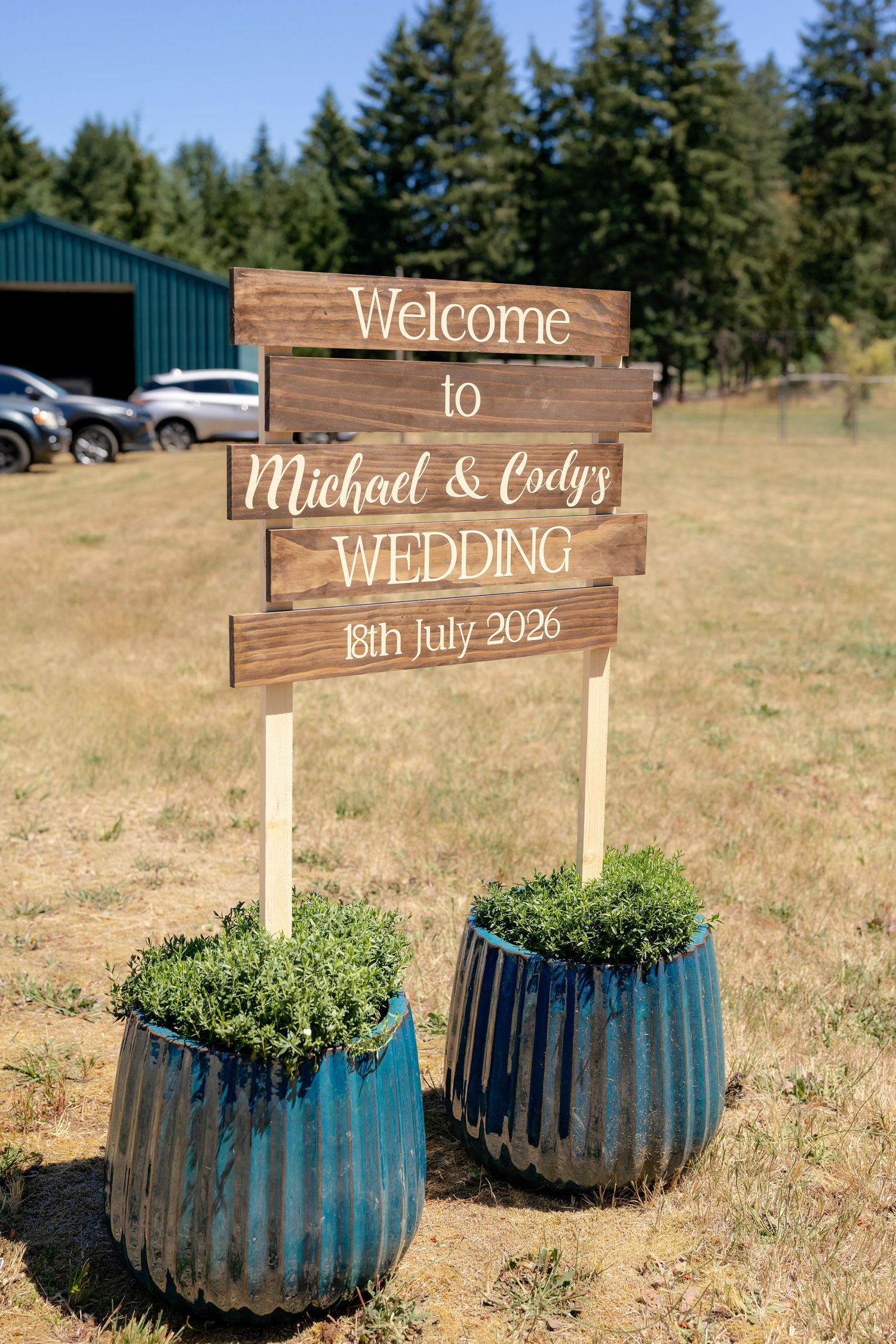 Rustic wooden welcome sign for Michael and Cody's wedding on 18th July 2026, displayed outdoors with blue corrugated metal planters.