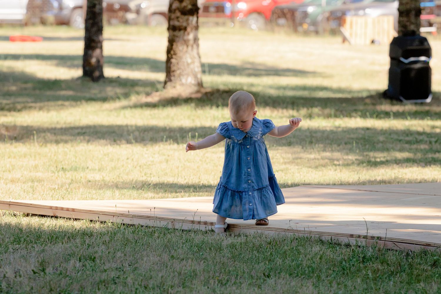 Toddler girl in blue denim dress walking on a wooden path in a sunny park with green grass.