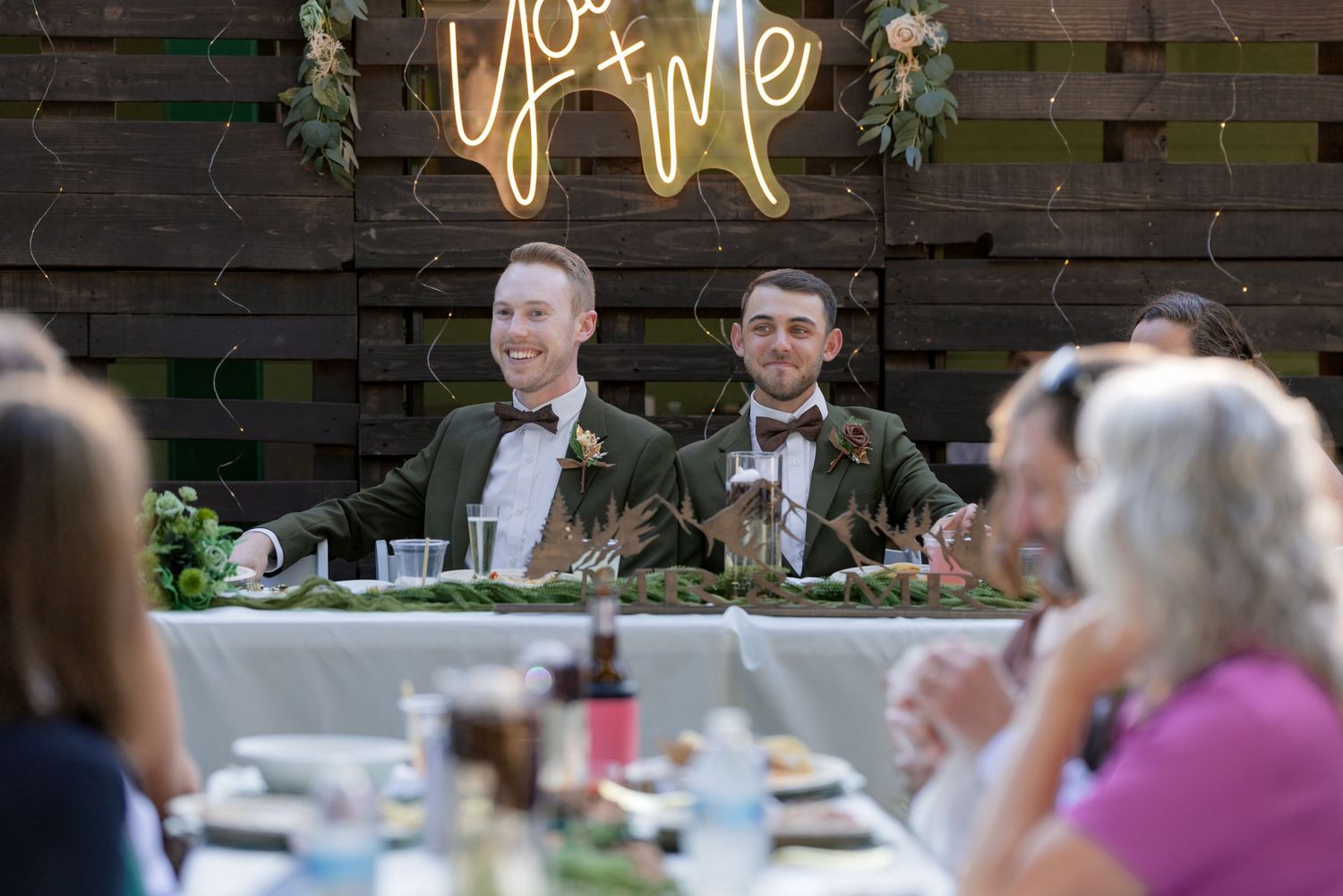 Two grooms in green suits smiling at sweetheart table under a You and Me neon sign at reception.
