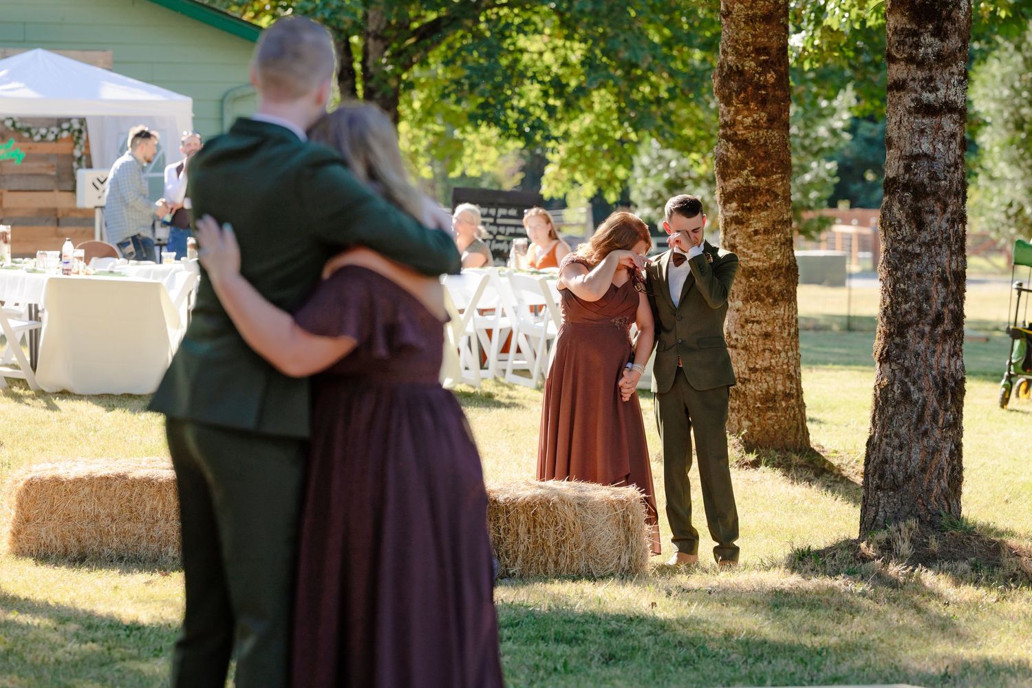 Two couples share emotional dances on grass at sunny outdoor rustic wedding reception ceremony.