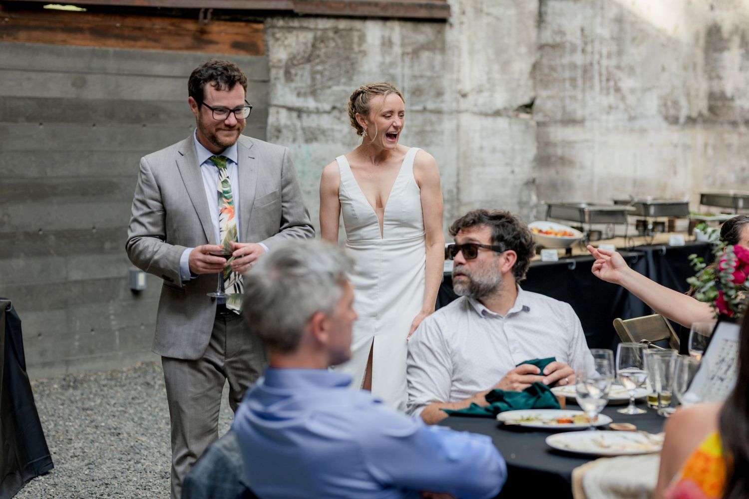 Bride and groom laugh with wedding guests during outdoor reception at rustic industrial venue with string lights.