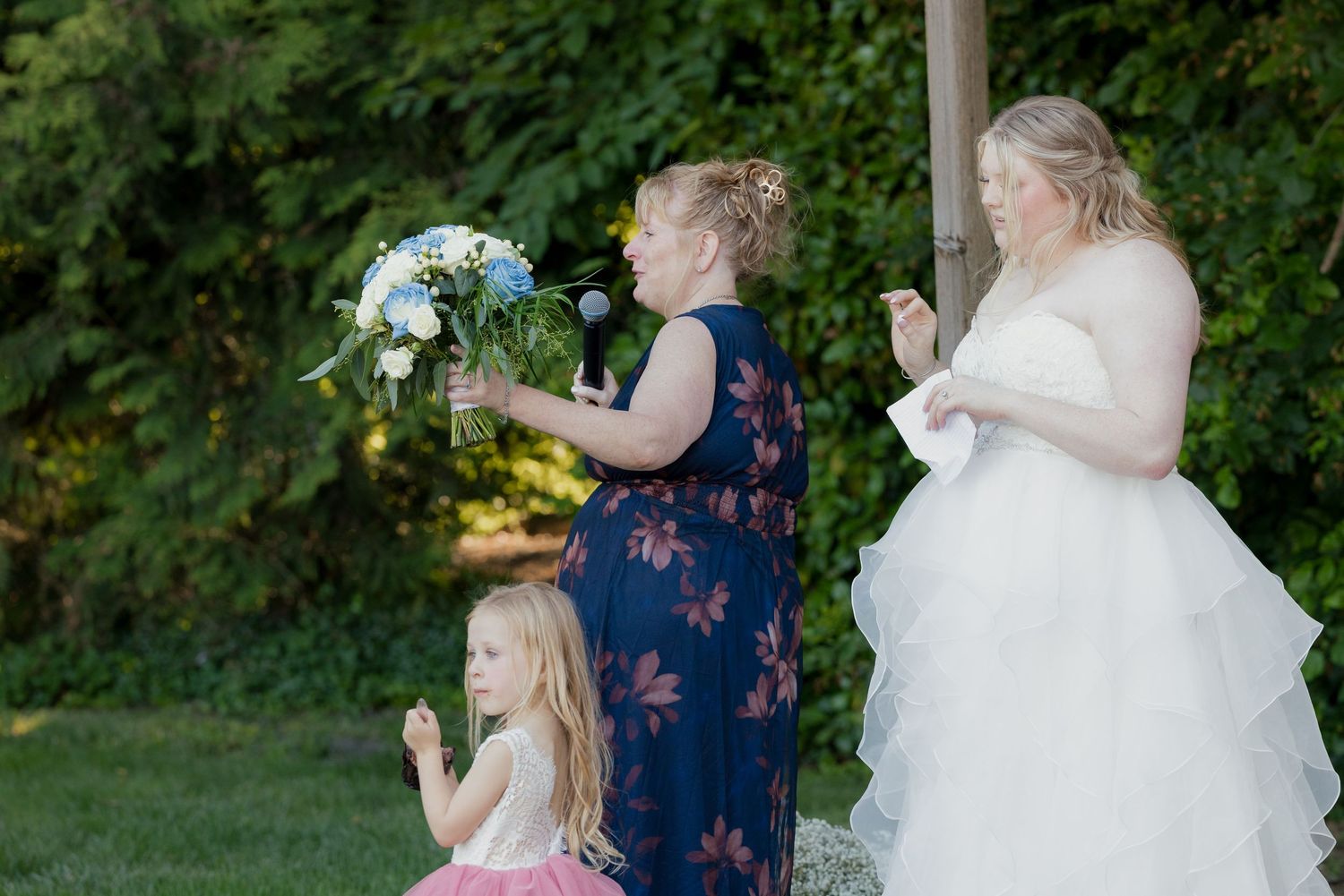 Bride in white gown receives bouquet from woman in navy floral dress as flower girl kneels nearby outdoors.