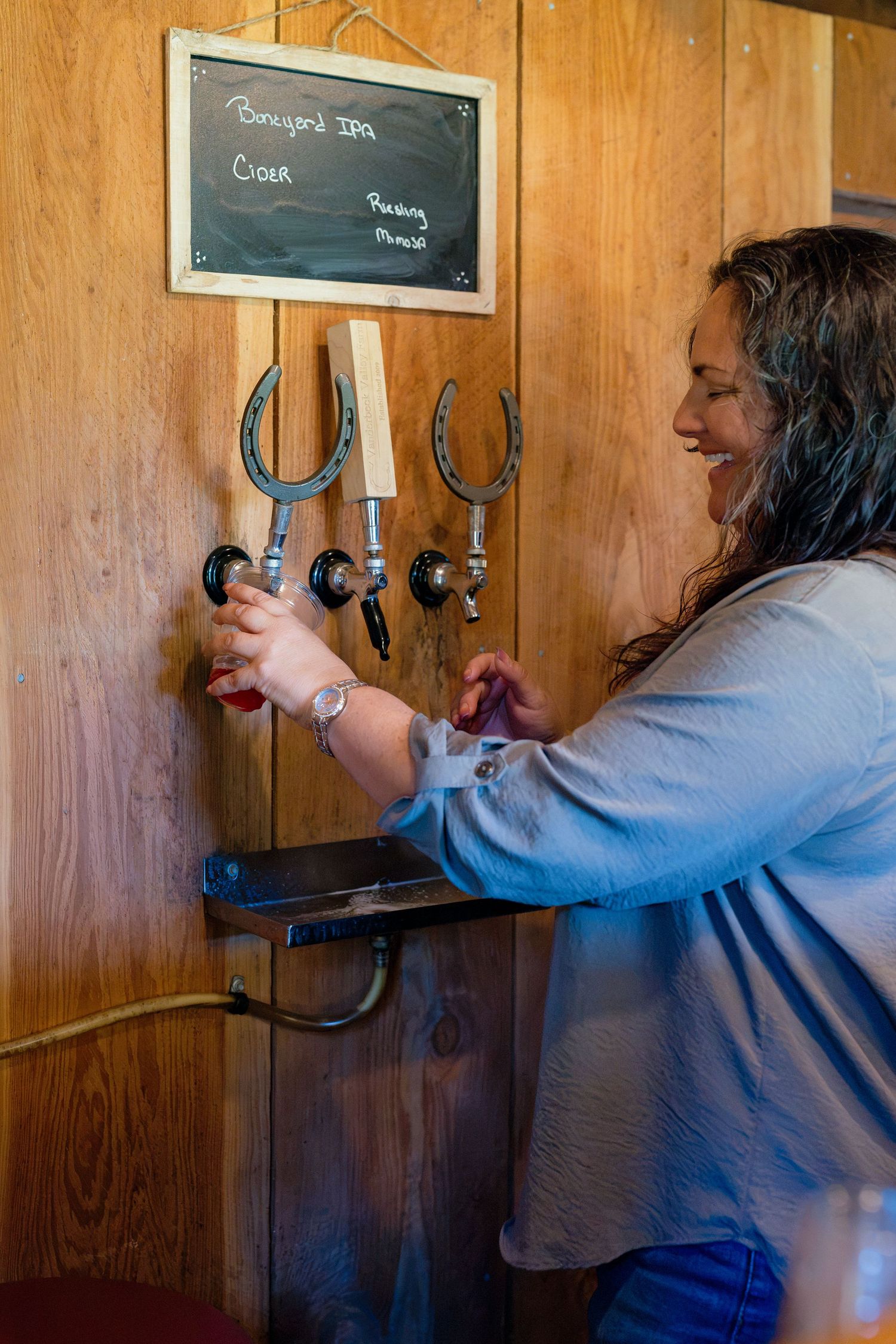 Smiling woman pouring draft beer from horseshoe tap handles at rustic wooden bar.