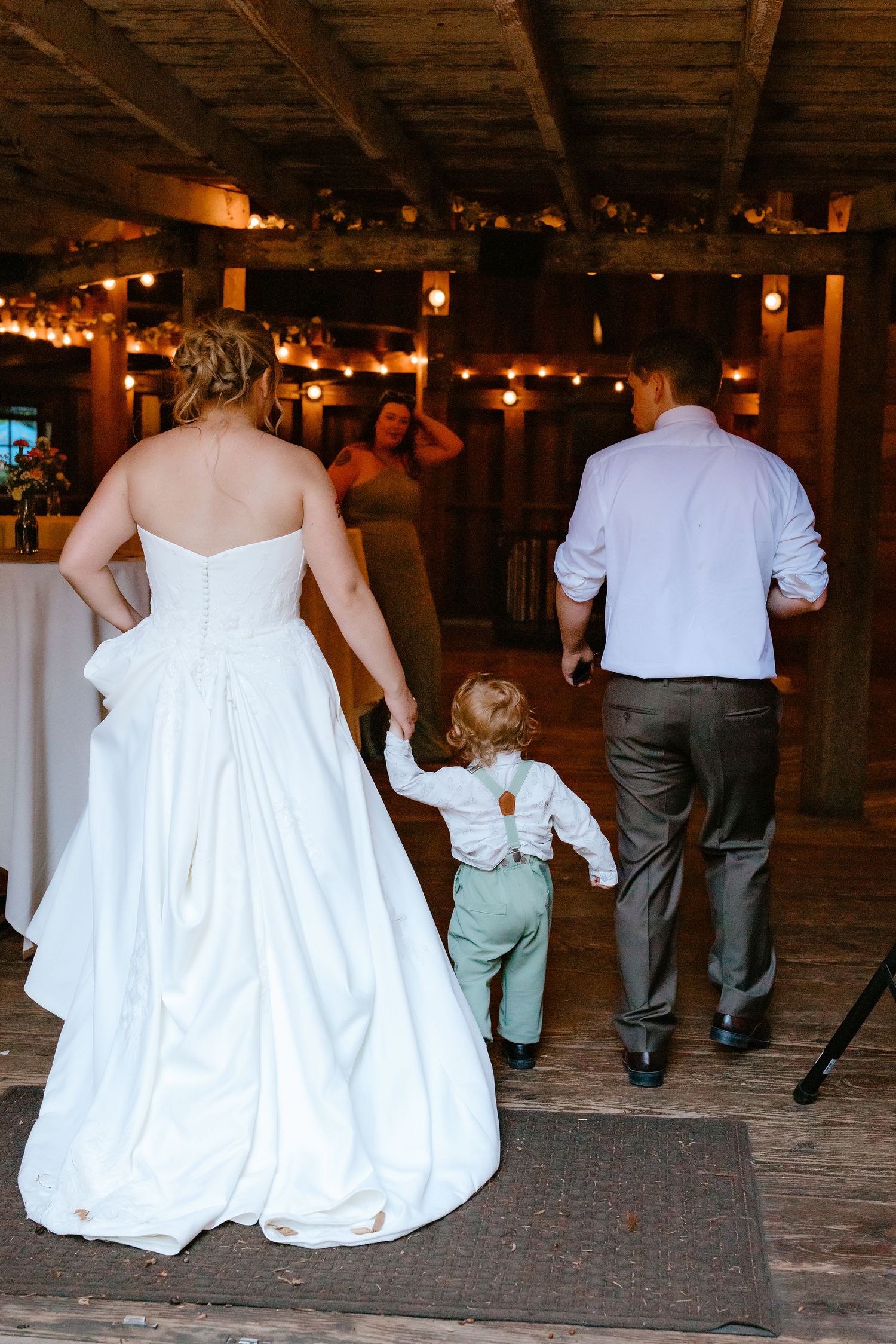 Bride and groom walk with young child in barn wedding reception.