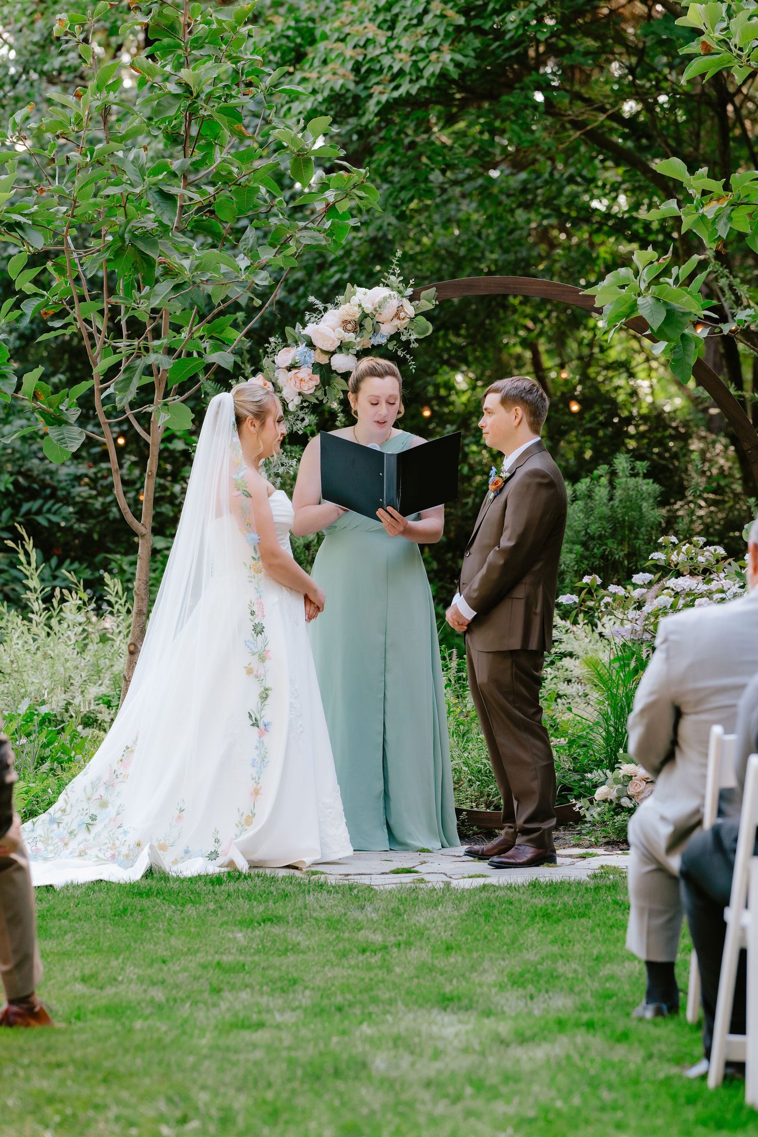Bride and groom exchange vows with officiant at elegant outdoor garden wedding ceremony under floral arch.
