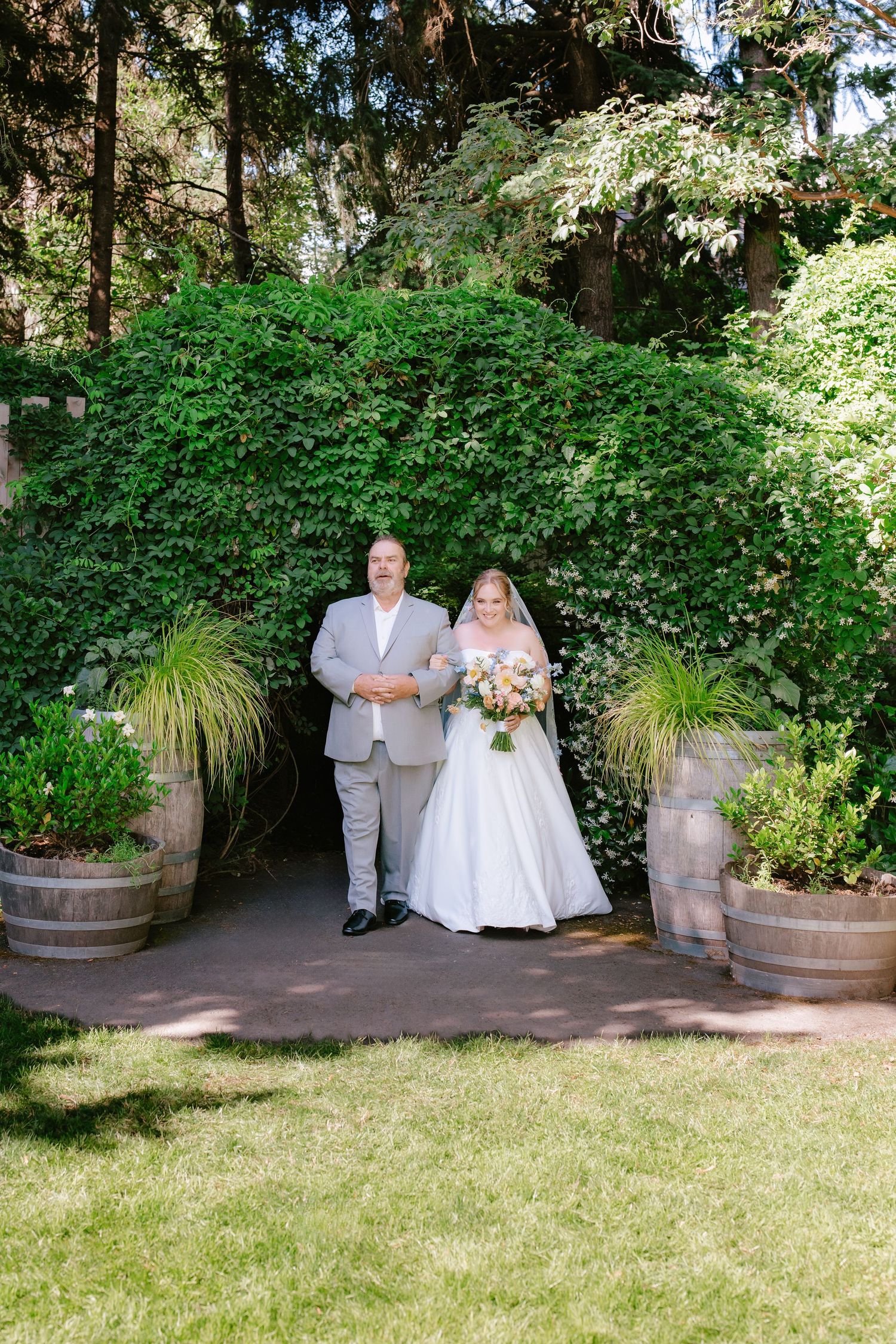 Bride in white gown with bouquet walks with escort through garden arch at outdoor wedding ceremony.