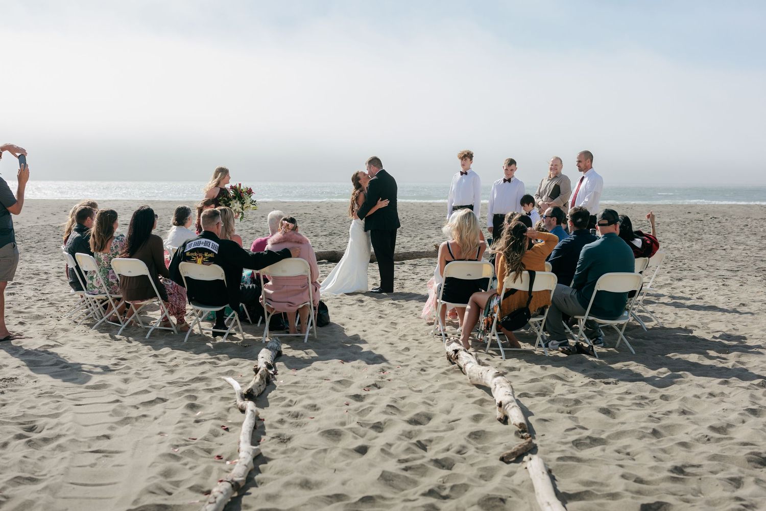 Wedding ceremony setup on a beach with guests seated in wooden chairs along rope-lined aisles.