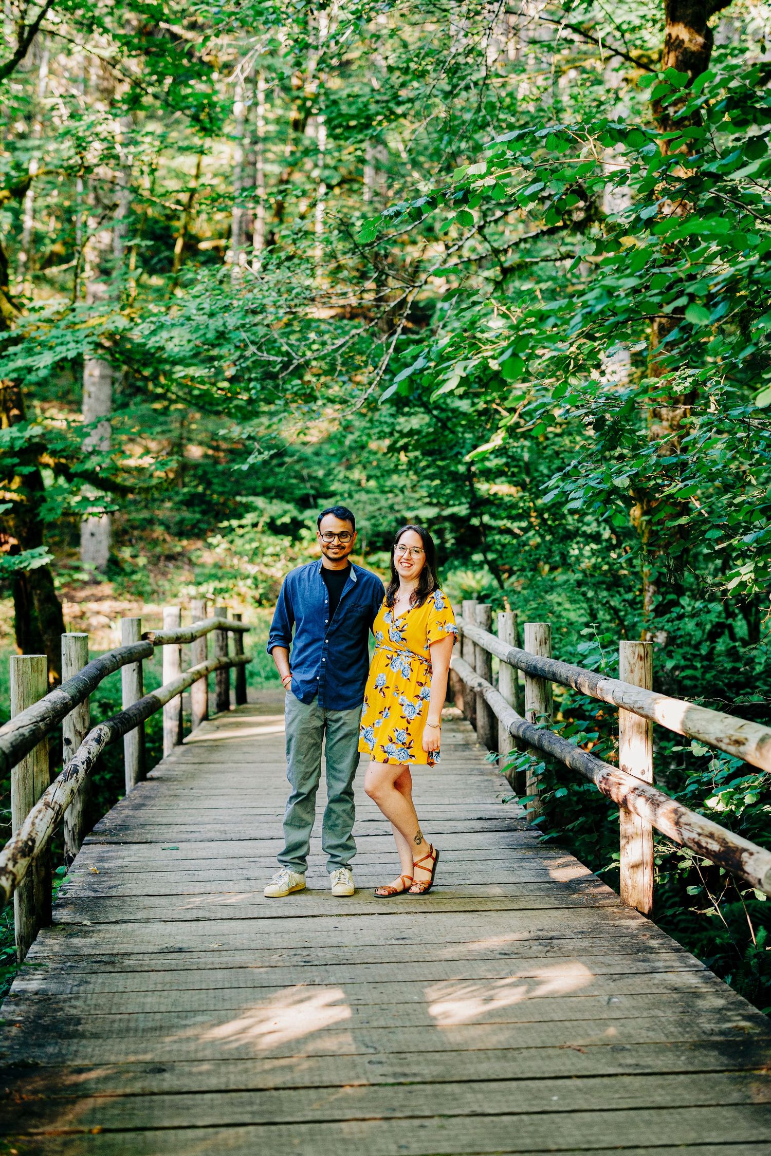 Couple standing together on a wooden boardwalk trail surrounded by lush green forest on a sunny day.
