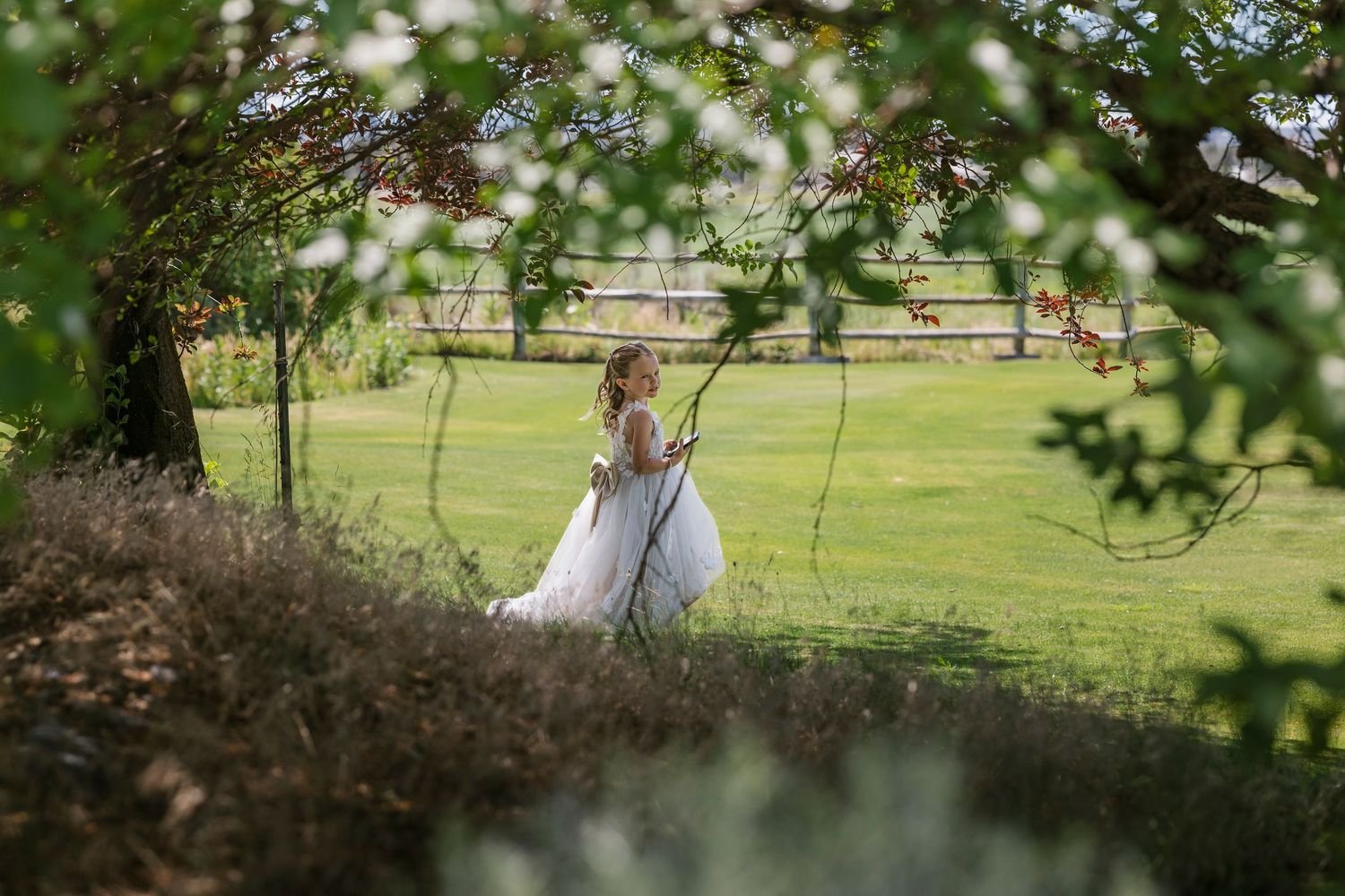 A bride in a white wedding gown sits on a rustic wooden swing in a sunlit garden setting.