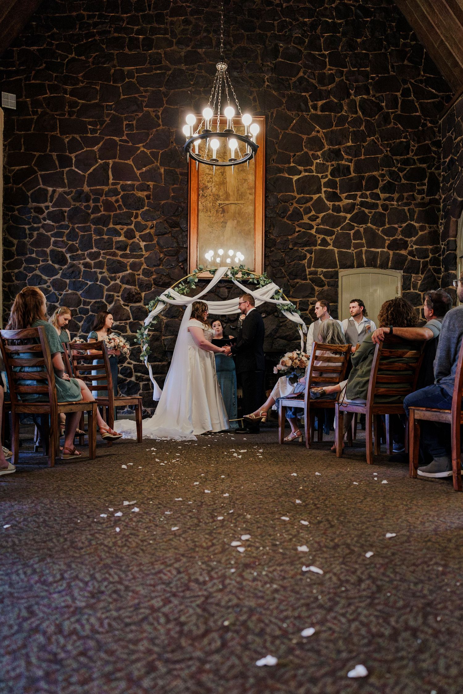 Wedding ceremony in rustic stone-walled venue with chandelier and scattered flower petals.