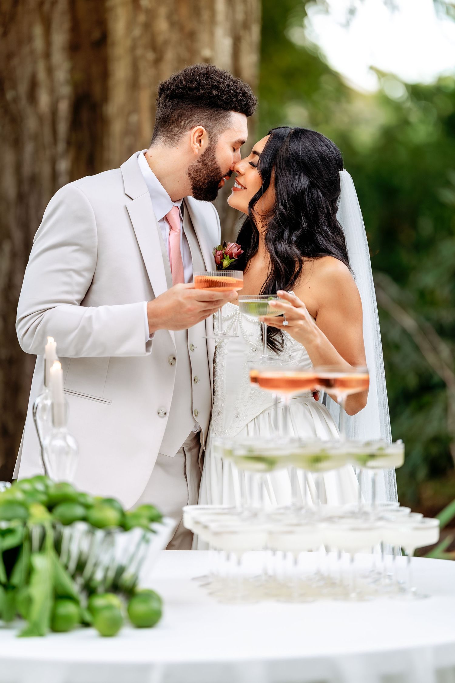 Newlyweds share a romantic moment while toasting with cocktails next to a champagne tower at their outdoor wedding reception.