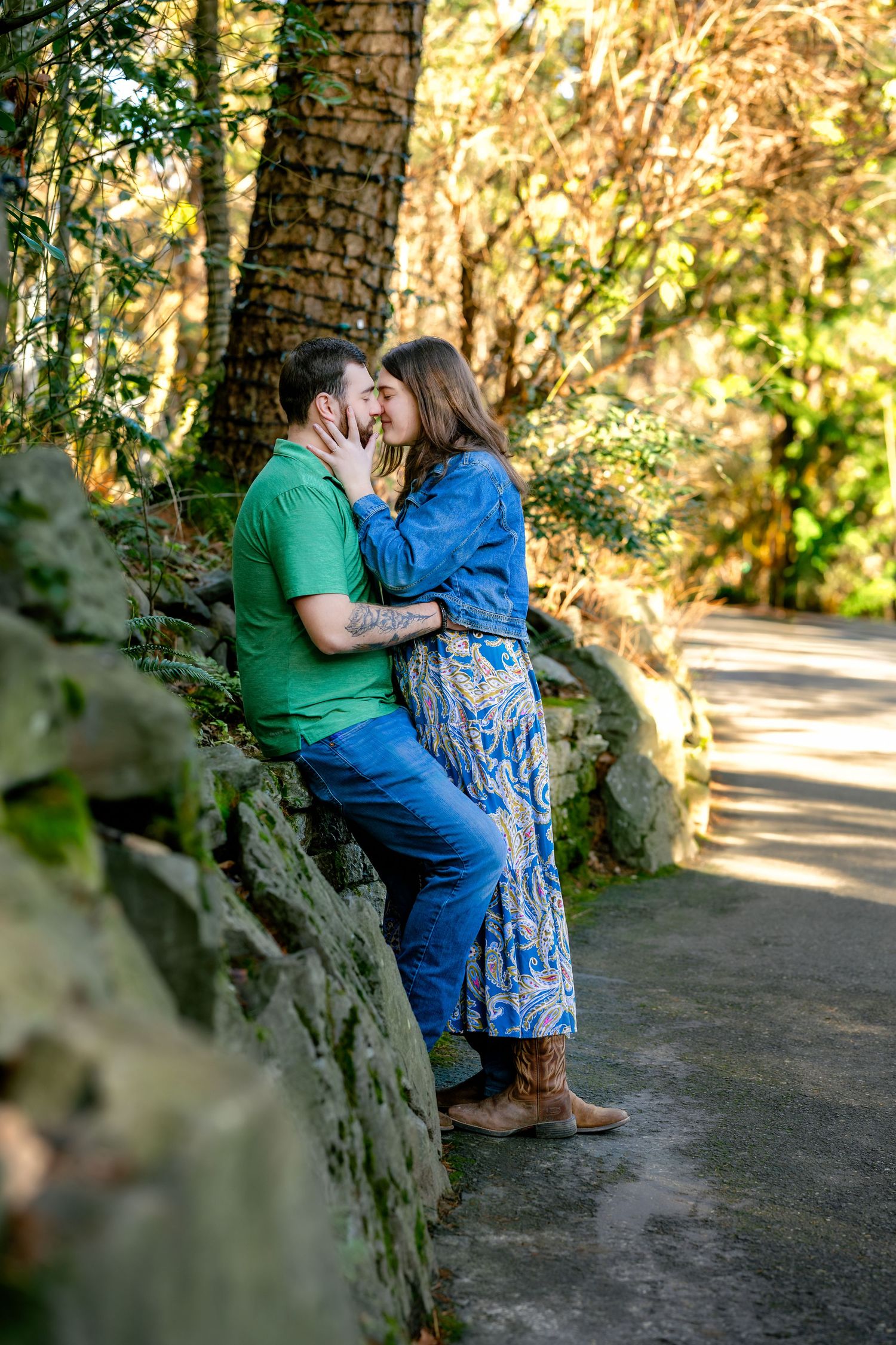 A romantic moment between a couple sitting on a stone wall in a park setting with dappled sunlight.