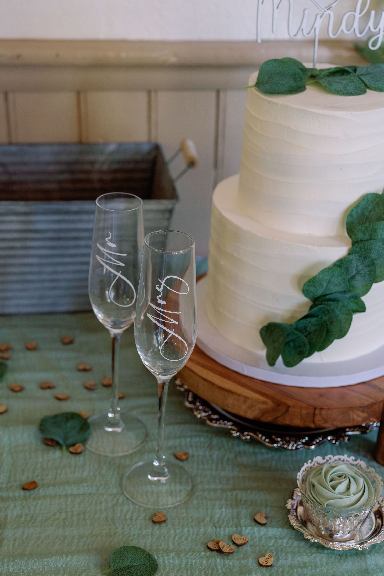 White wedding cake with green eucalyptus leaves and champagne flutes on a mint tablecloth.