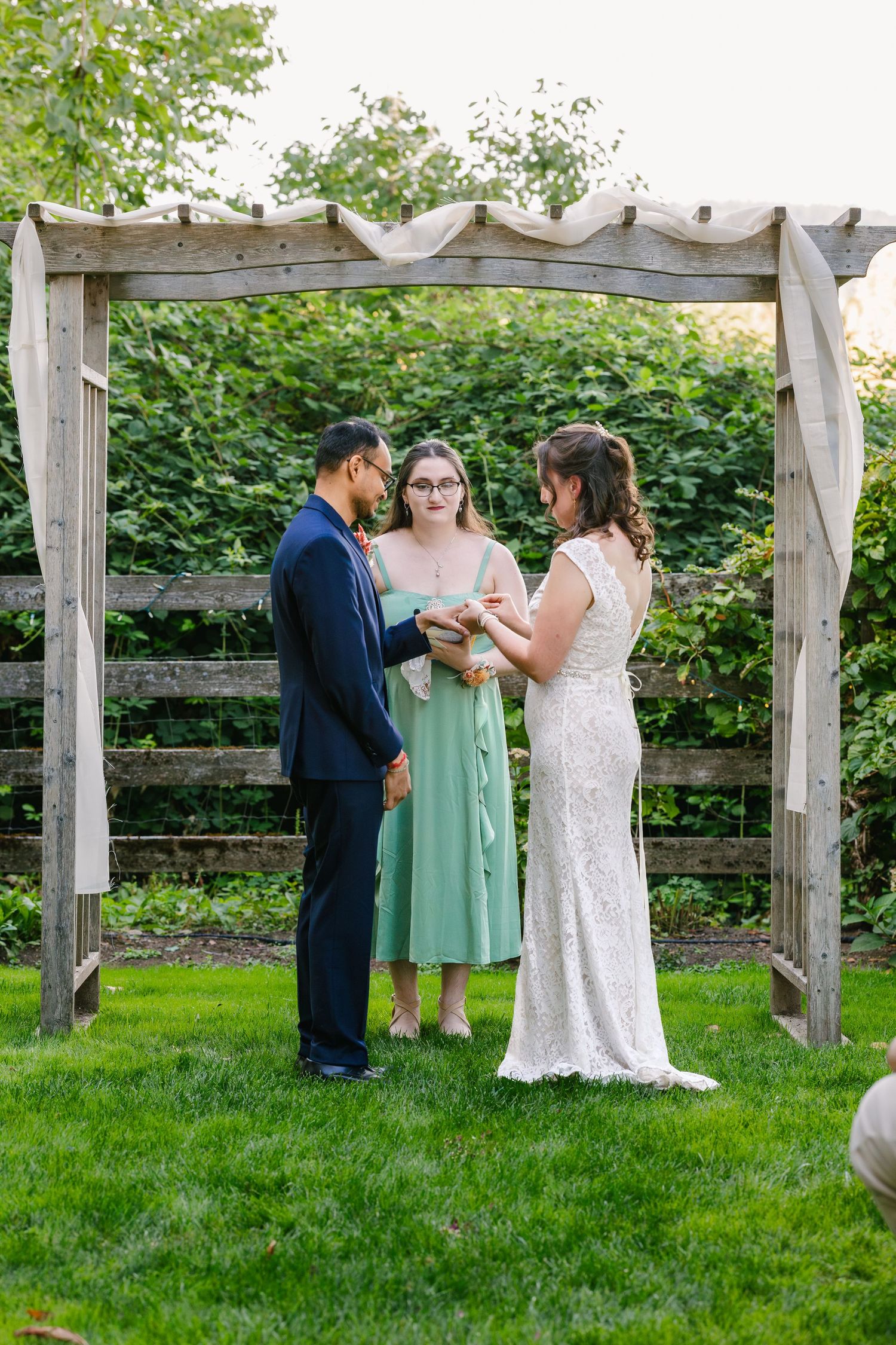 Wedding party gathers on lush green lawn near wooden pergola during outdoor celebration.