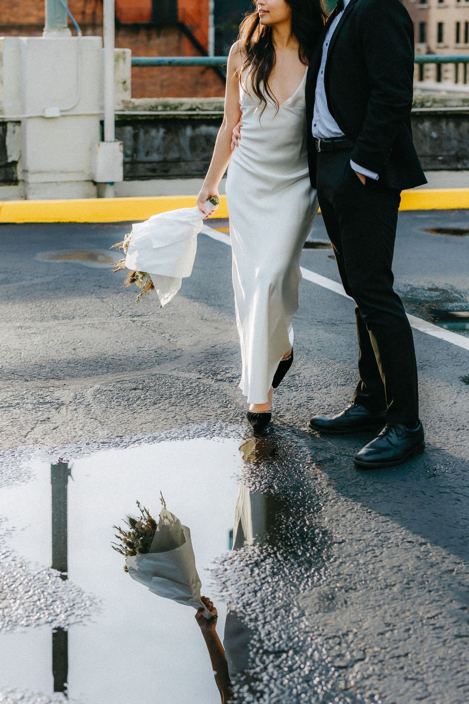 Close-up of formal shoes and dress hem reflected in puddle on city street.
