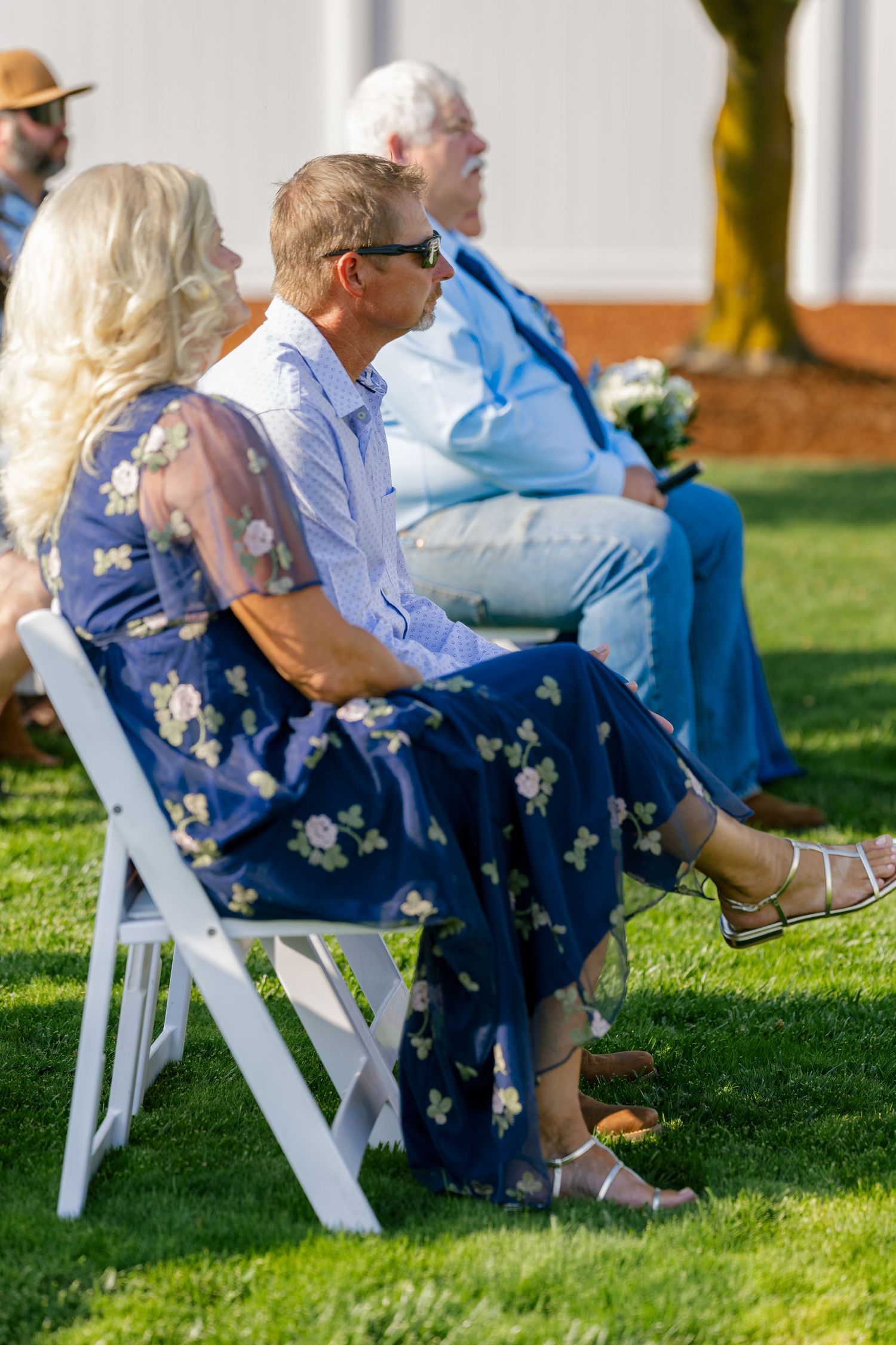Wedding guests seated in white chairs on a sunny lawn wearing blue floral dresses.