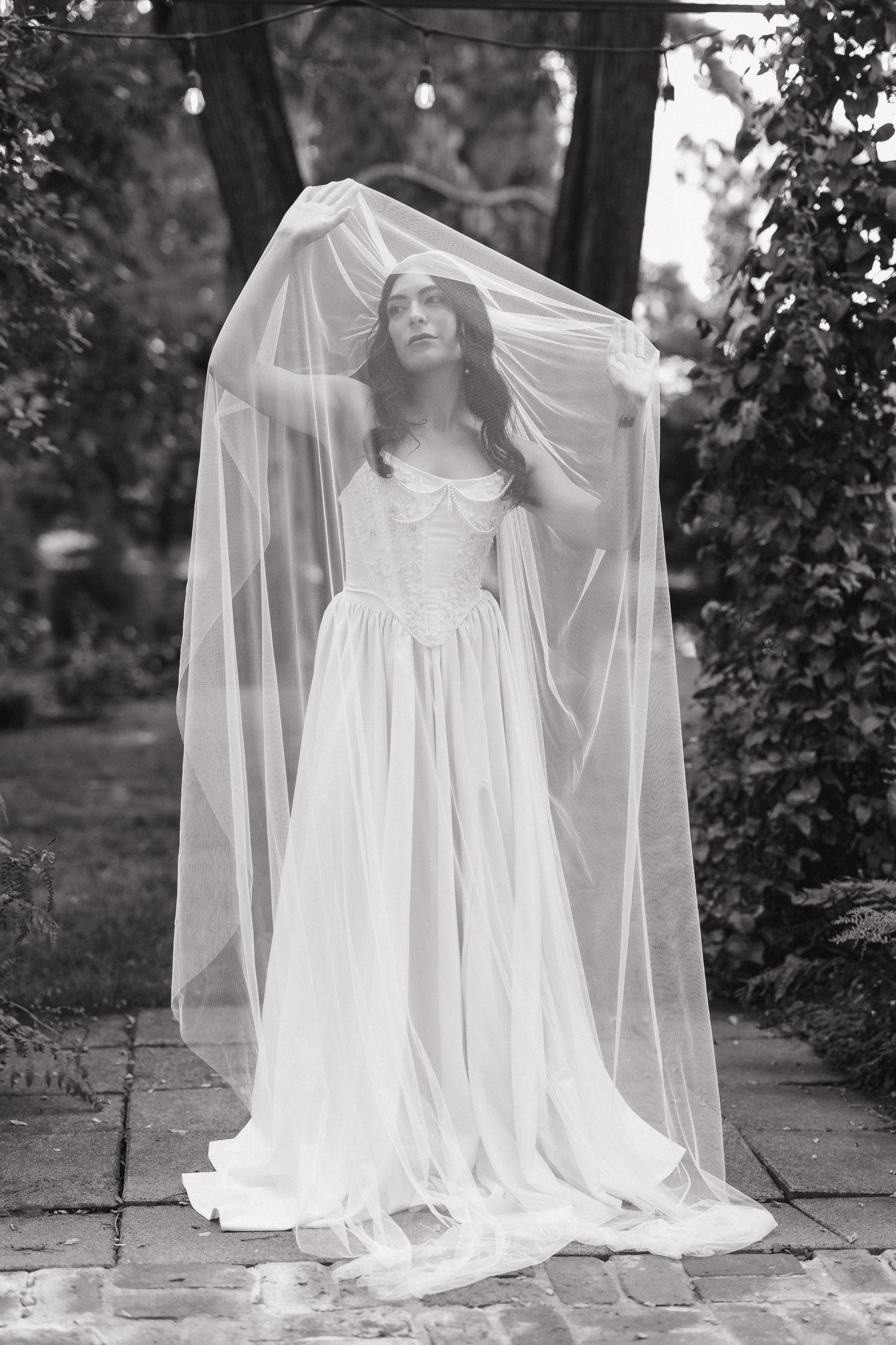Black and white portrait of a bride in a flowing veil standing on stone pavement.