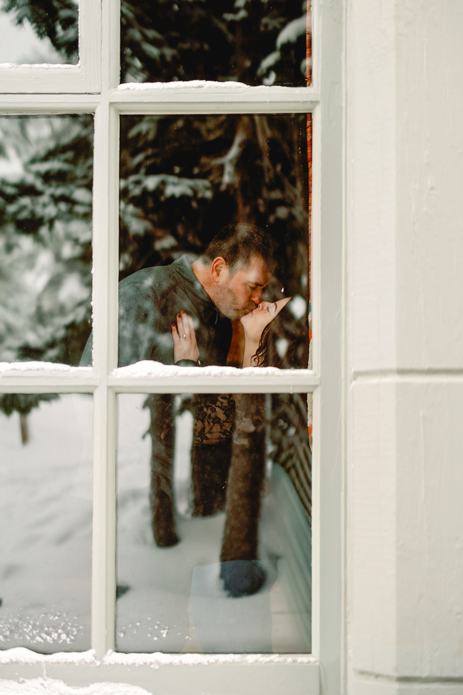 Two people playfully peek through a white-framed window on a snowy winter day.