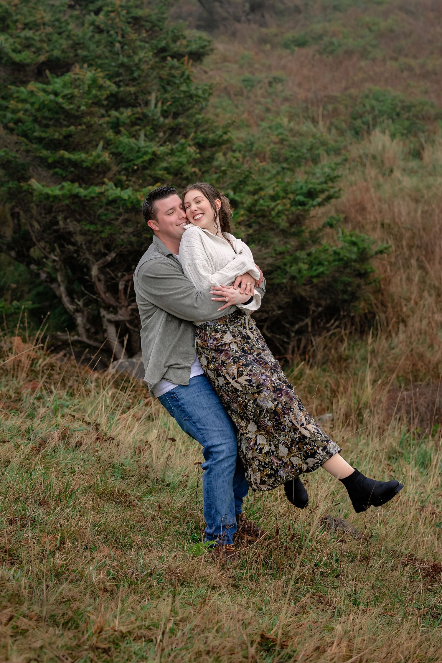 A romantic couple shares playful dips and embraces on a grassy hillside surrounded by evergreen trees.