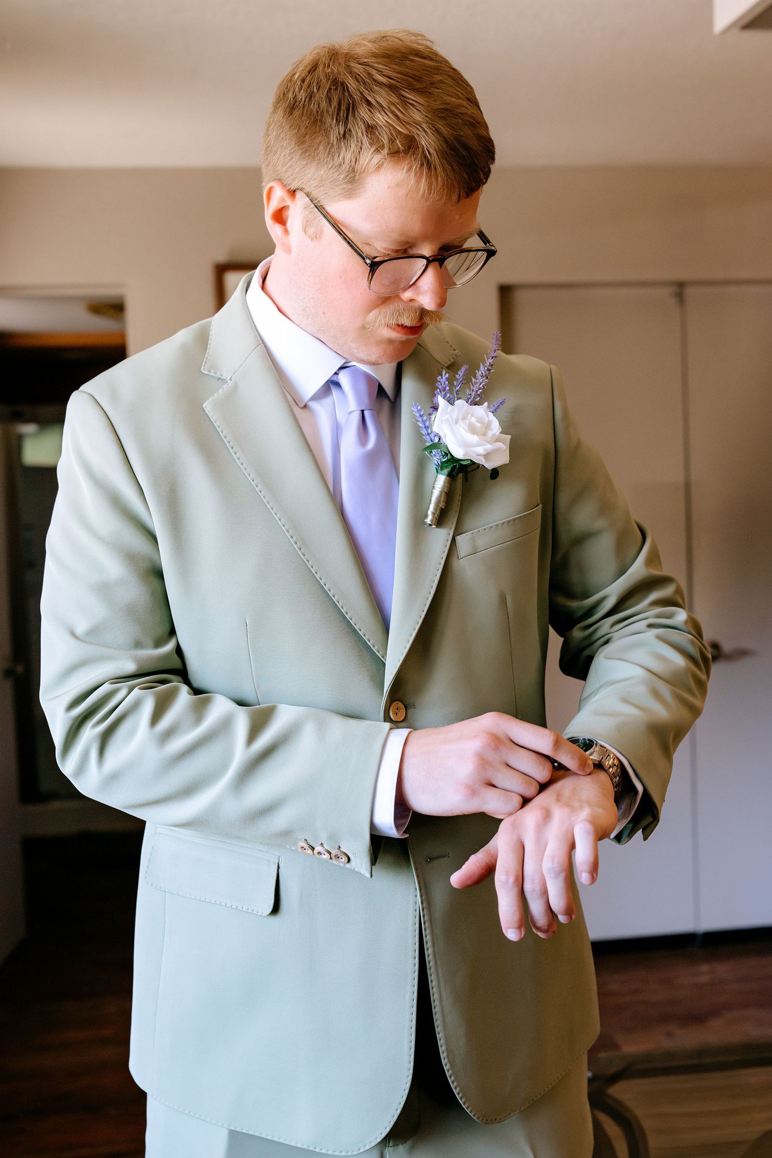 Groom in sage green suit with lavender tie and boutonniere checks his watch while getting ready for wedding.