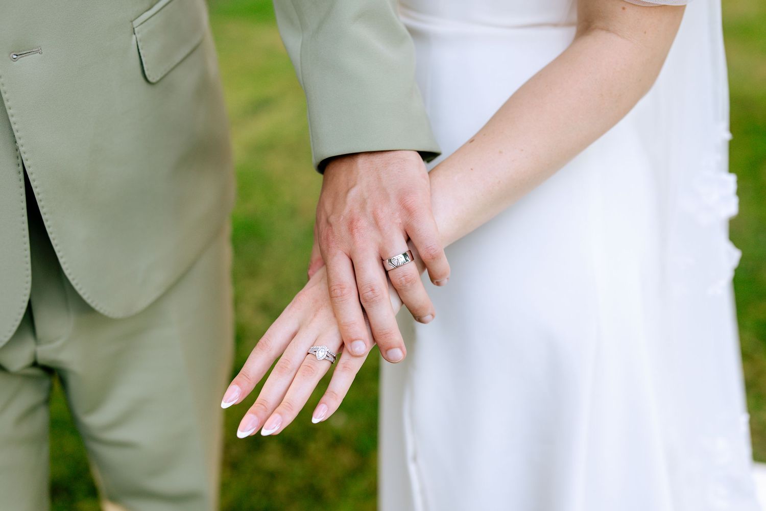 Close-up of bride and groom displaying their wedding rings, wearing sage suit and white wedding gown.