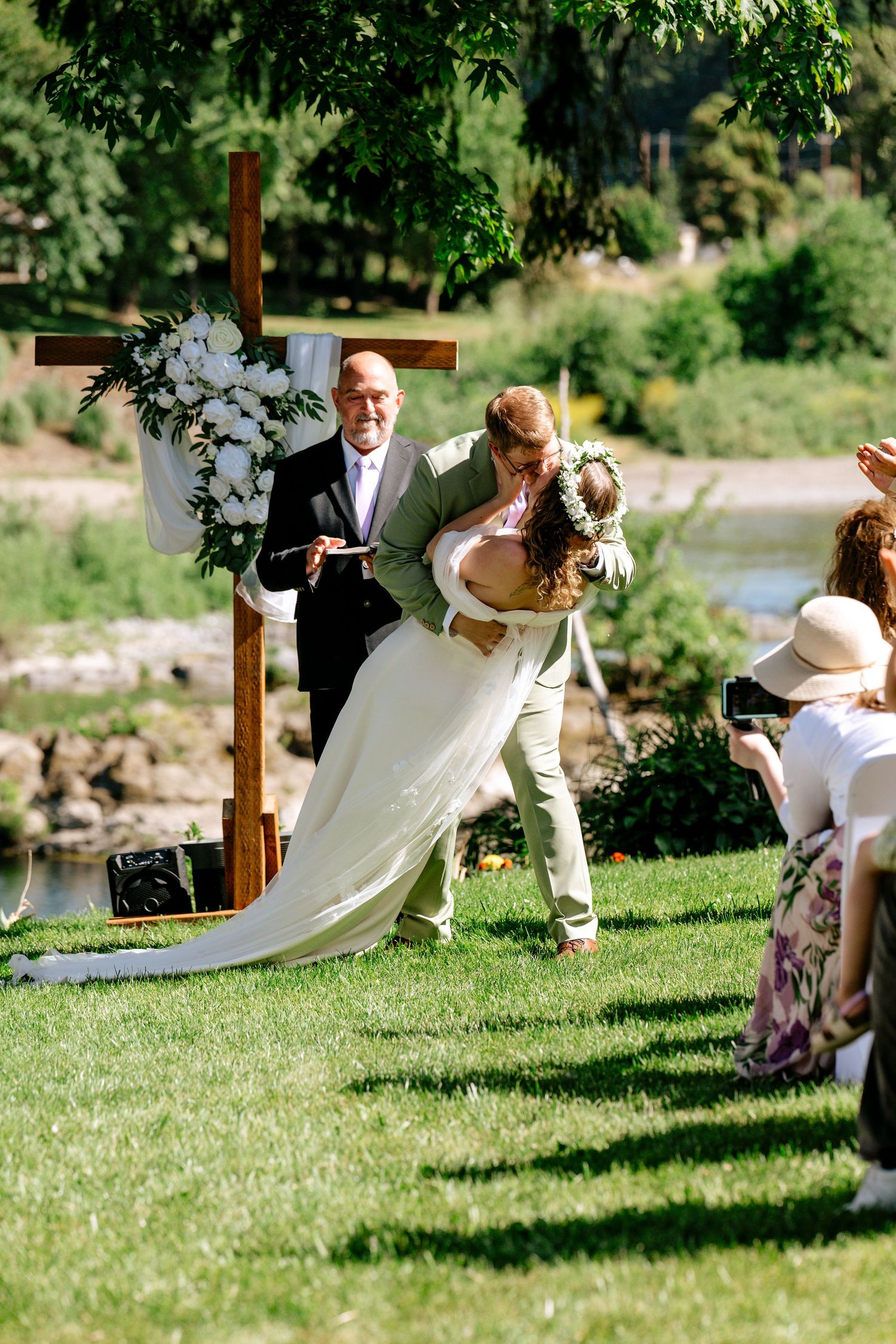 Groom dips bride for a kiss at outdoor wedding ceremony beside wooden cross altar on green lawn.
