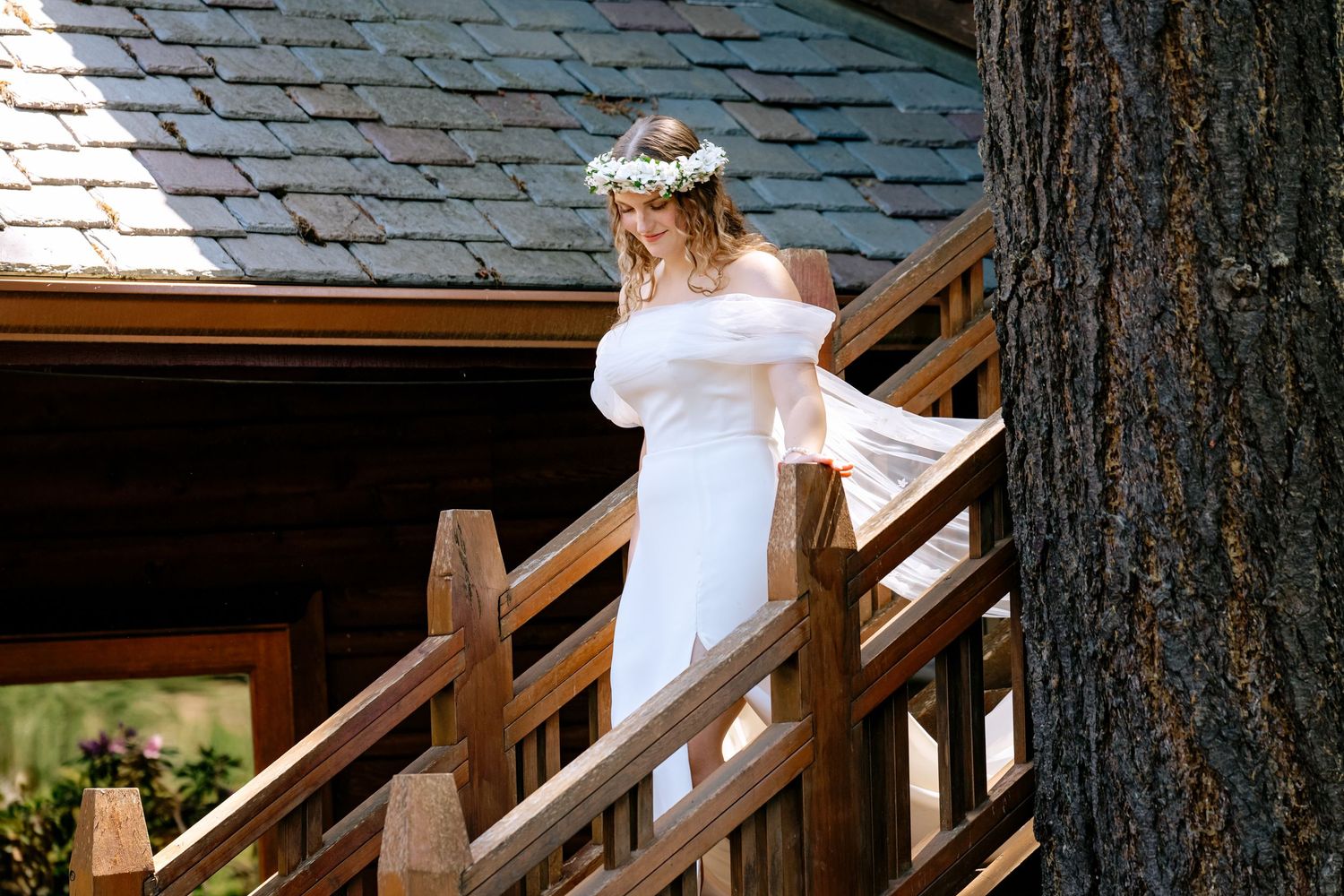 Bride in white off-shoulder gown and floral crown stands on wooden stairs beside rustic cabin and tree.