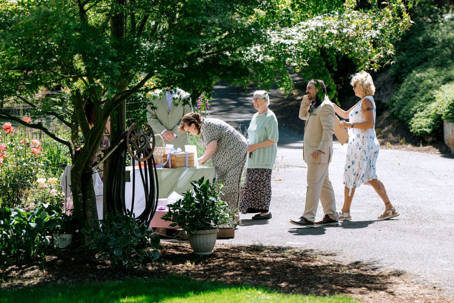 Wedding guests arriving and greeting each other along a tree-lined garden path on a bright sunny day.