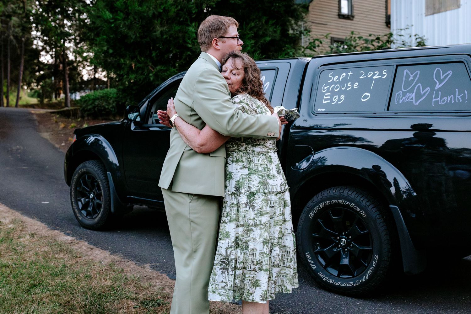 Groom in sage suit embraces a woman in floral dress beside a black truck with wedding date written on window.