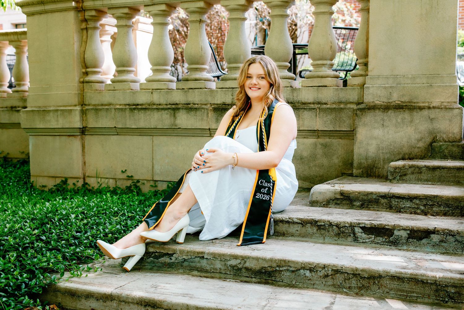 Smiling graduate in white dress and green stole sits on stone steps holding diploma at outdoor ceremony.