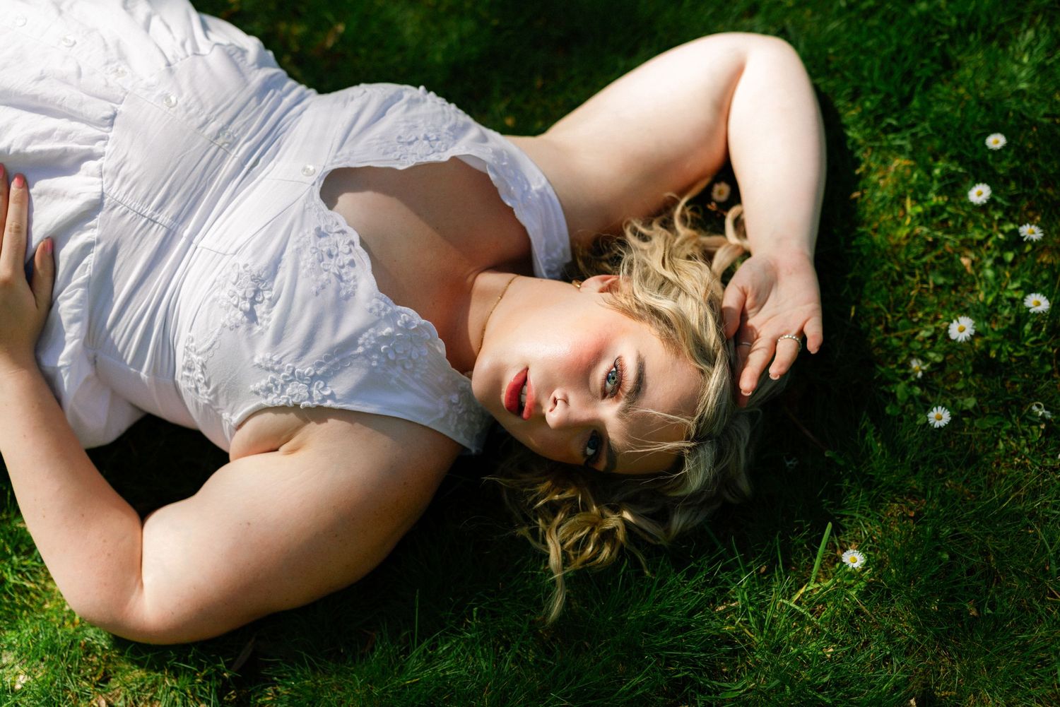Blonde woman in white dress lying on green grass with daisies, looking up in natural outdoor setting.