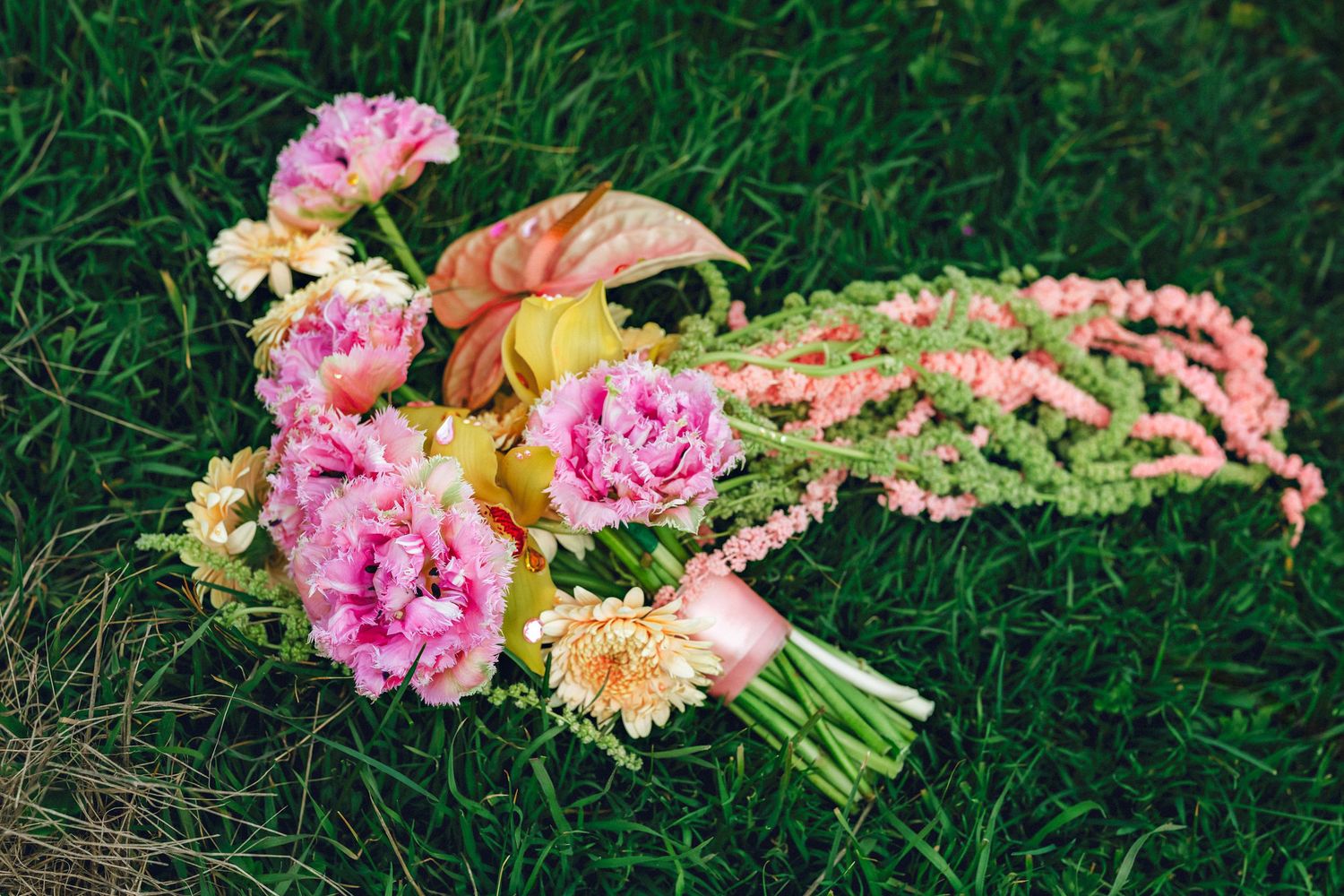 Bridal bouquet of pink peonies, yellow flowers, and coral anthurium lying on green grass.