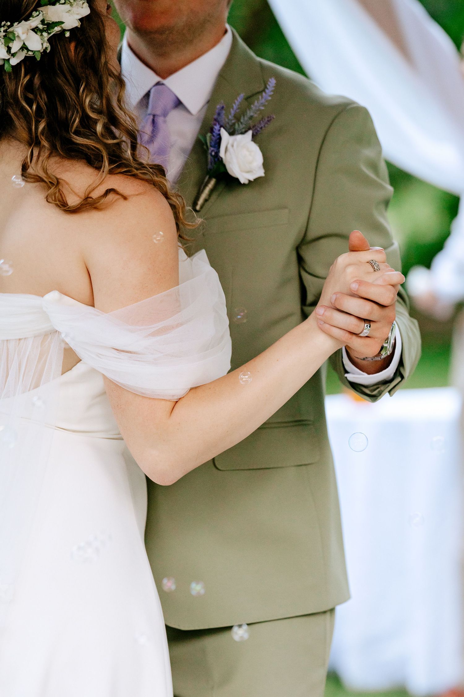 Bride and groom sharing first dance outdoors, she in white off-shoulder gown, he in sage green suit with floral boutonniere.