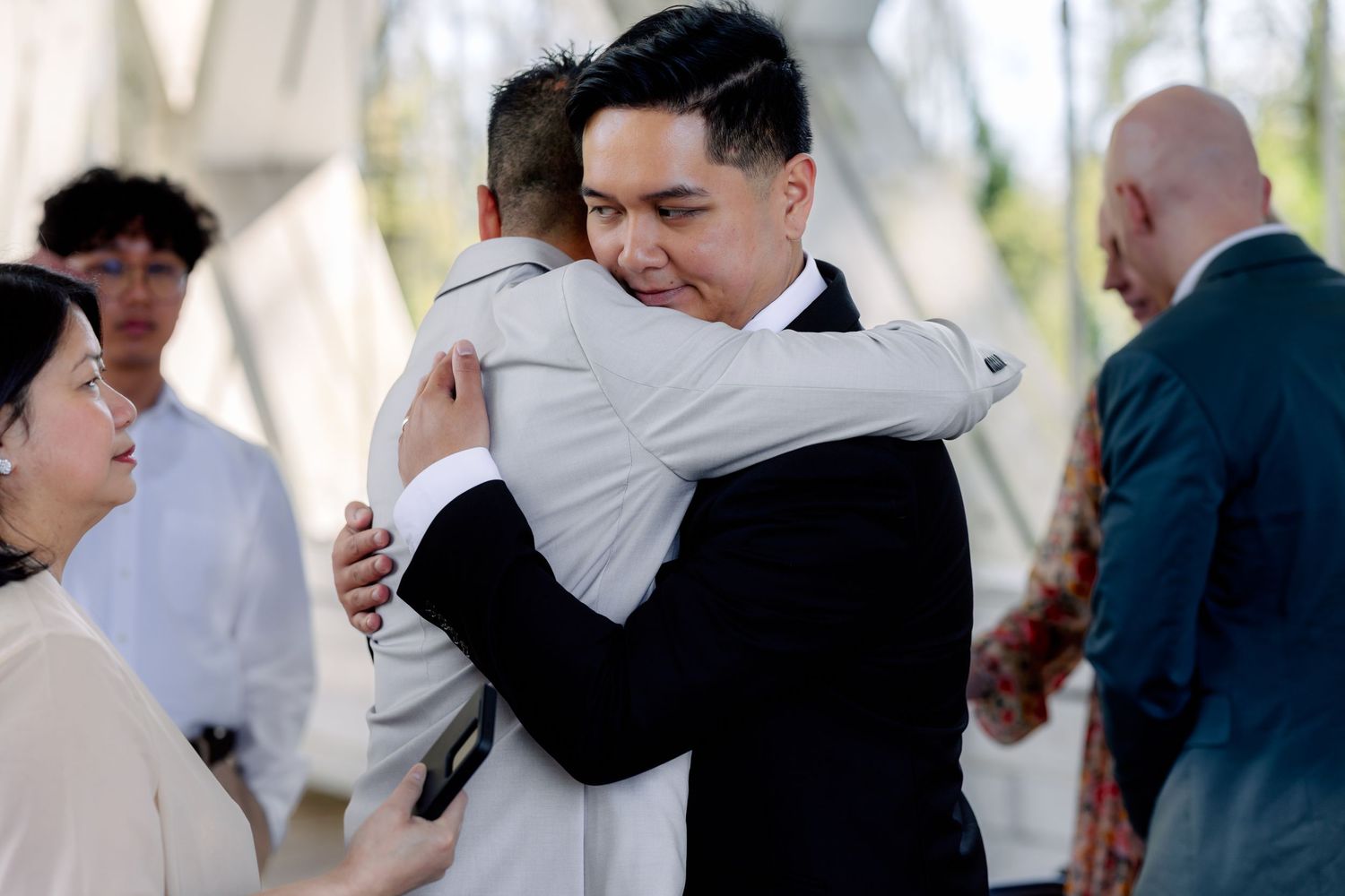 Two men in suits sharing an emotional hug at a wedding celebration outdoors.