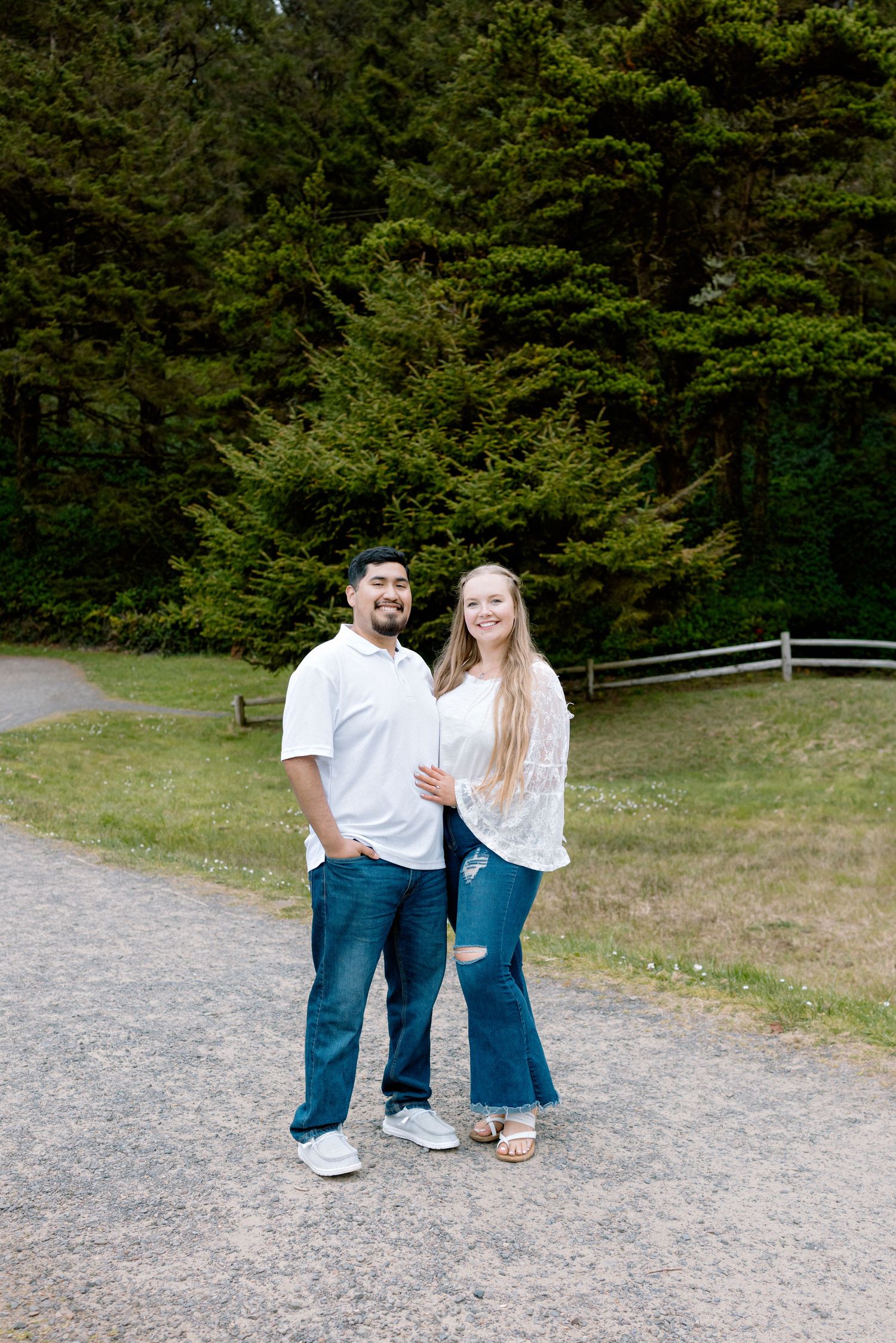 Smiling couple in casual white tops and jeans posing together on a gravel path surrounded by evergreen trees.