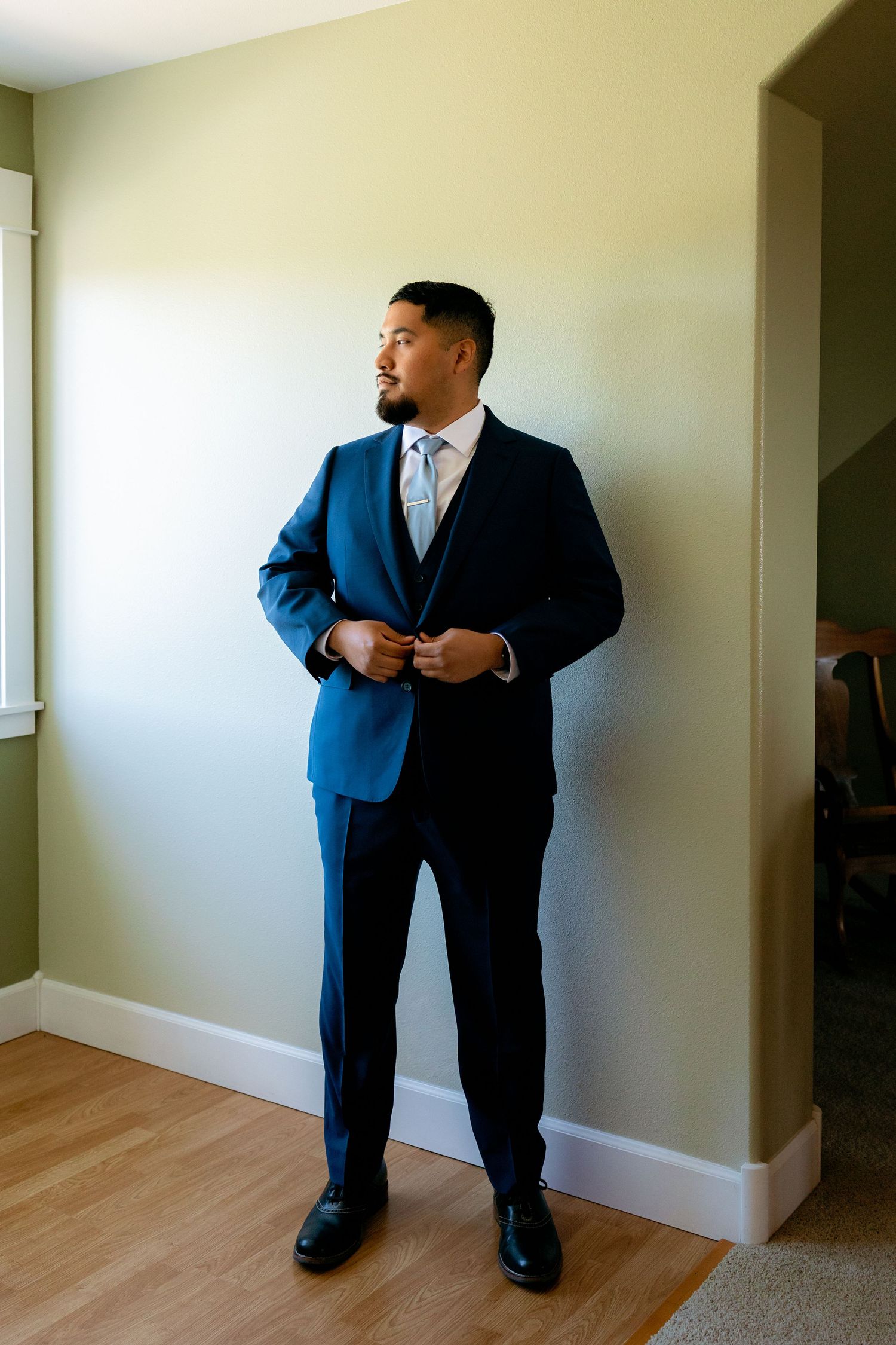 Groom in navy blue suit buttoning jacket while standing in room with green walls before wedding ceremony.