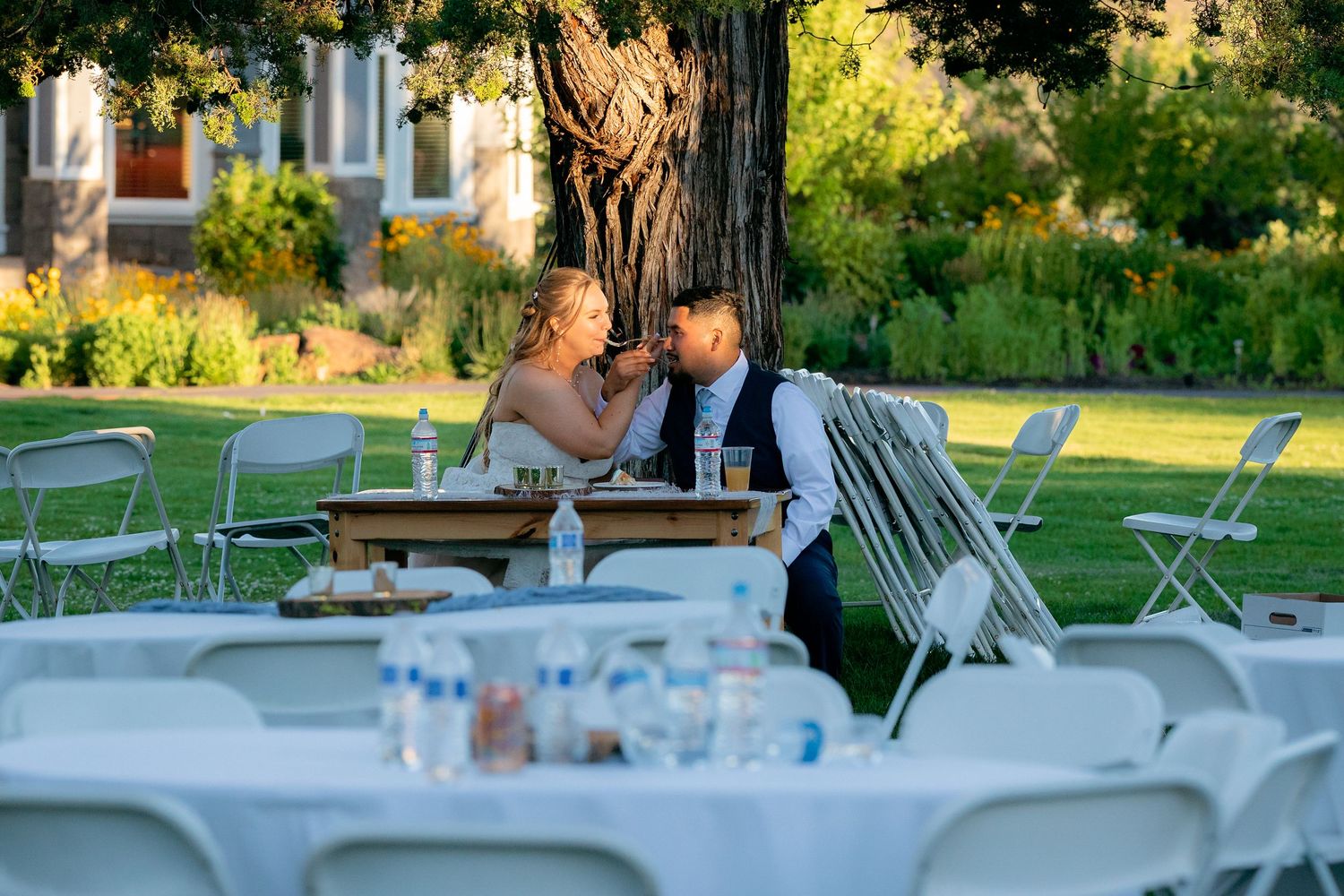 Newlywed couple shares intimate moment seated together at outdoor wedding reception on lush green lawn.