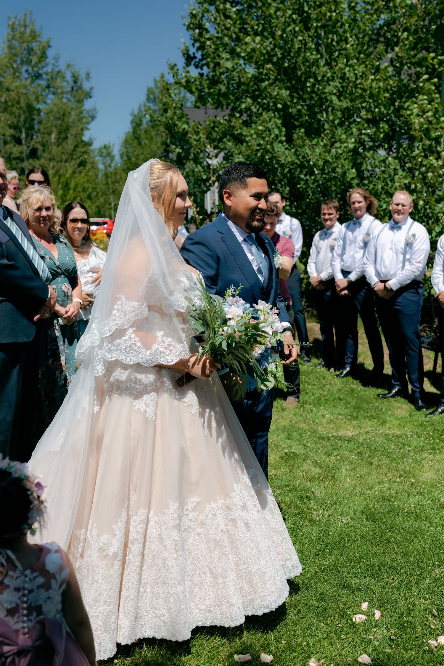 Bride and groom walking down outdoor aisle surrounded by smiling guests on a sunny summer wedding day.
