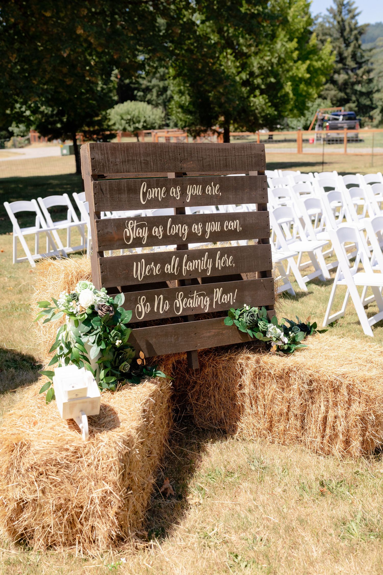 Rustic wooden pallet wedding sign on hay bales reads Come as you are, no seating plan, with floral decor.