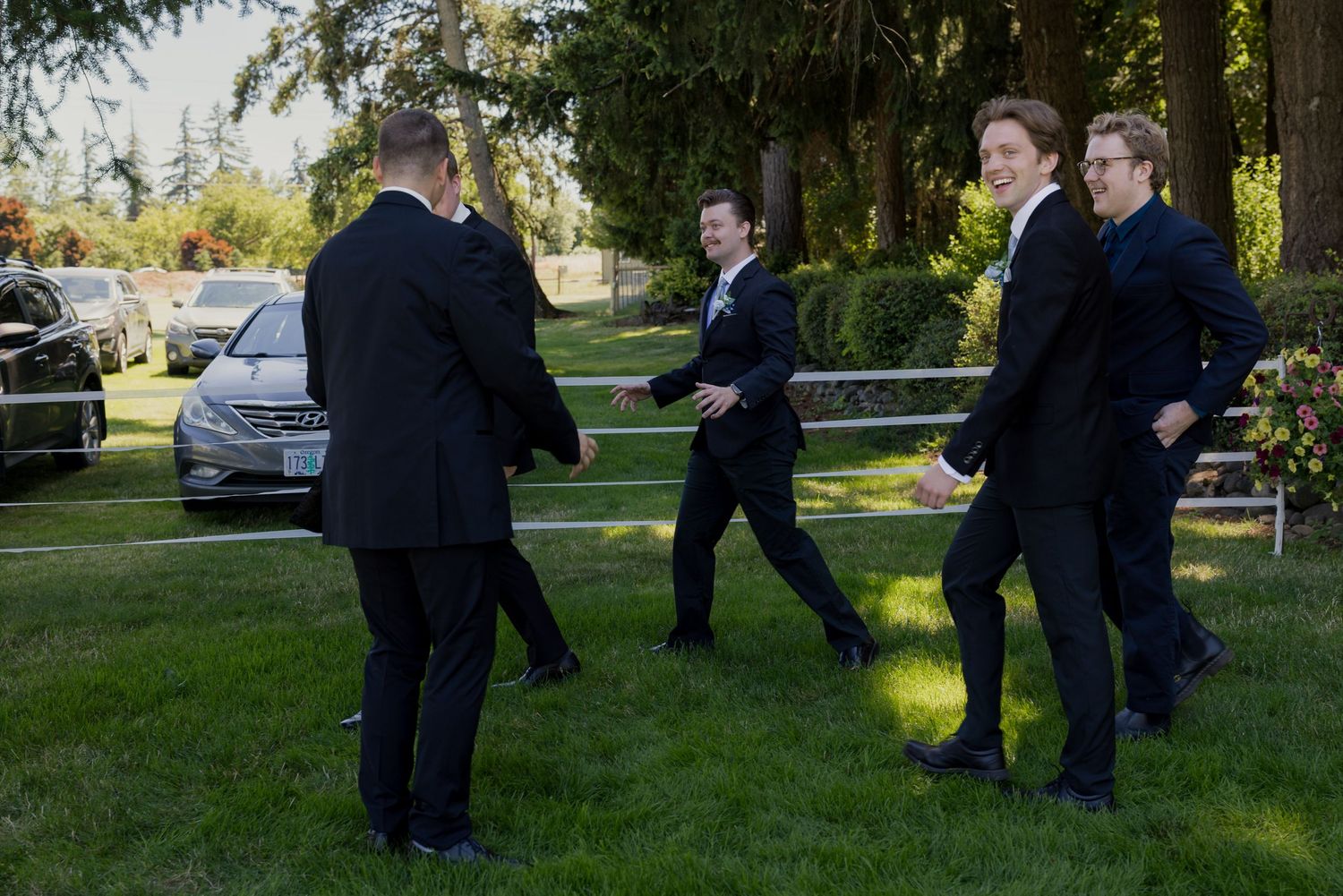 Group of young men in dark suits walking on grass at an outdoor formal event or wedding ceremony.