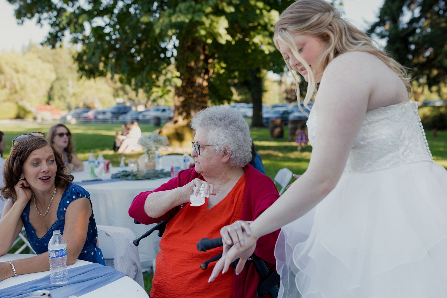 Bride in white gown sharing a tender moment with elderly grandmother in wheelchair at wedding.