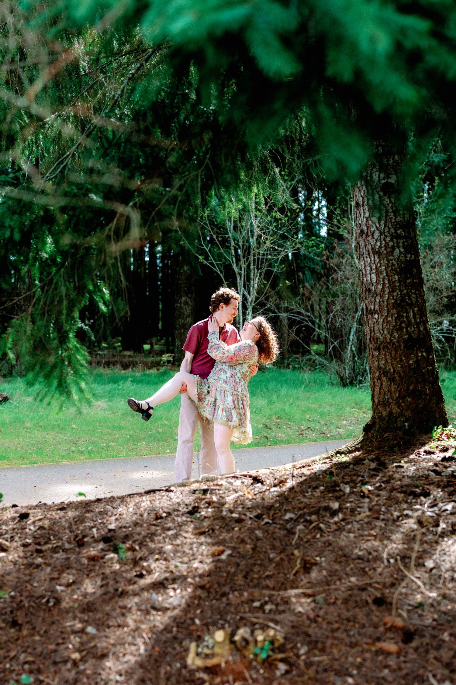 A couple shares a romantic dip pose under a lush green tree in a park setting.