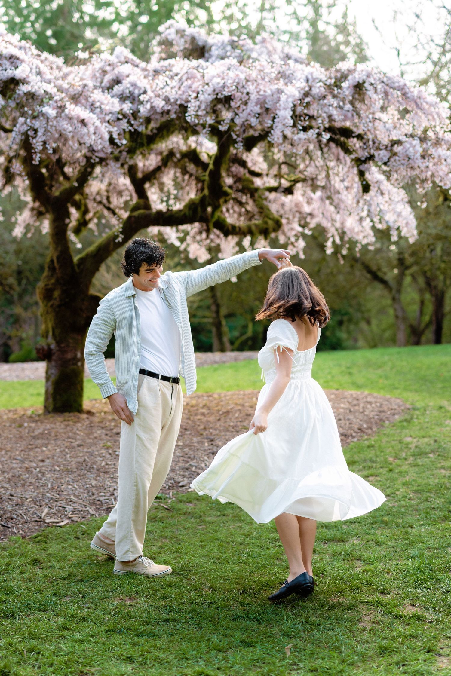 A couple dances playfully under blooming cherry blossom trees in a springtime garden setting.